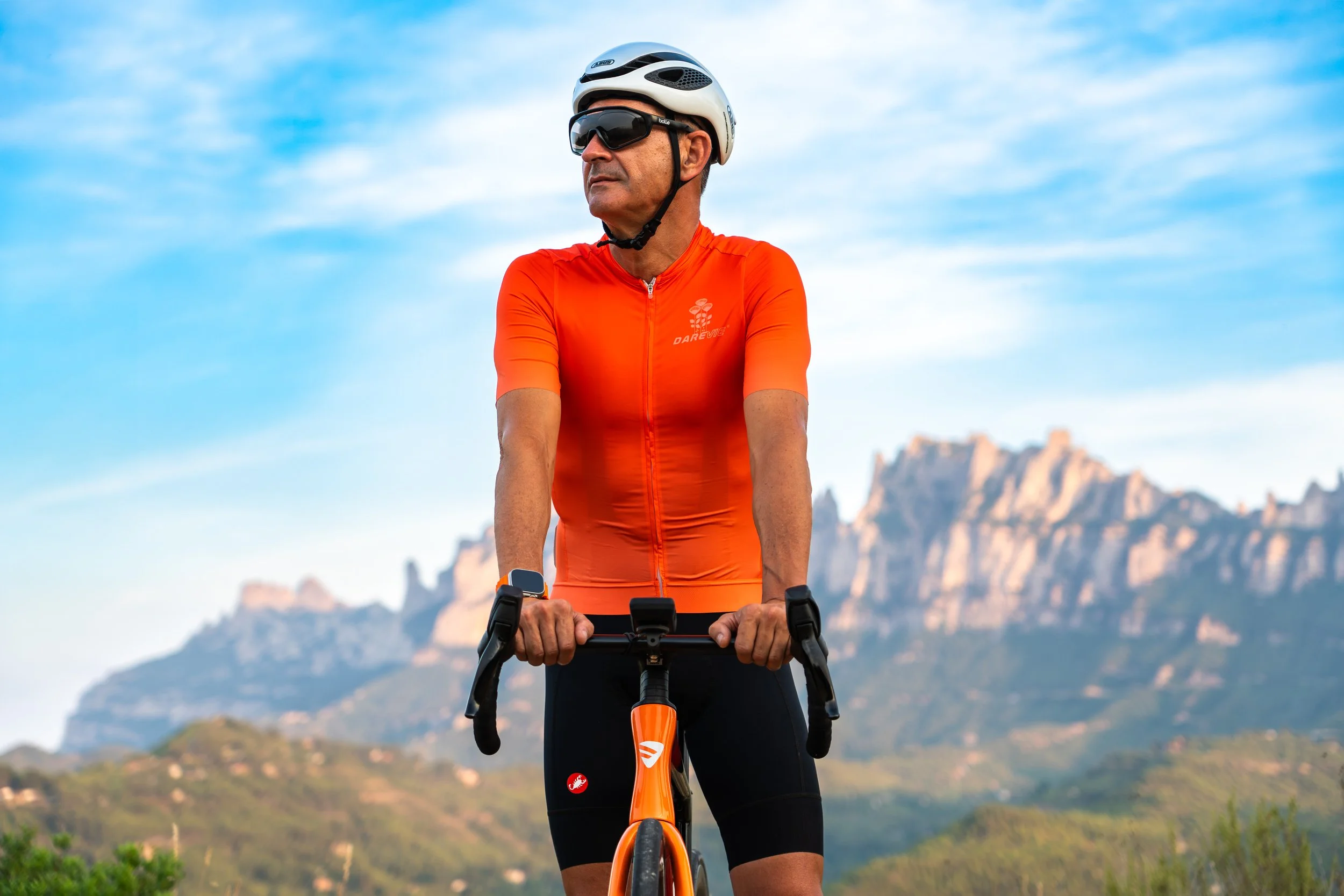 Un ciclista de sexo masculino en un uniforme naranja, con casco y gafas de sol, montando una bicicleta en un paisaje montañoso con cielo despejado.