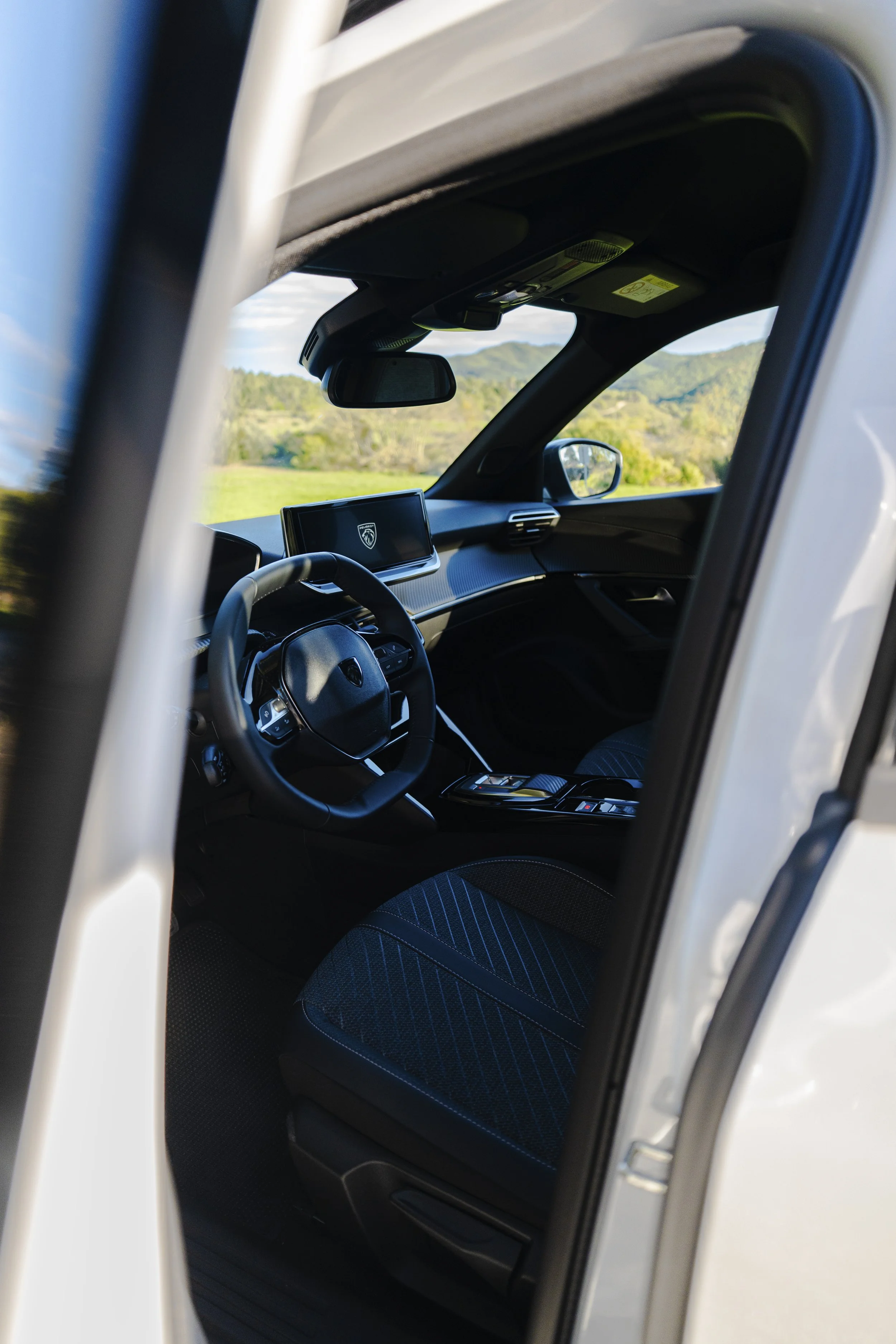 Interior de un vehículo, se aprecia el volante, la pantalla de infoentretenimiento con el logotipo de Peugeot, el espejo retrovisor y paisaje de naturaleza con montañas y árboles por la ventana.