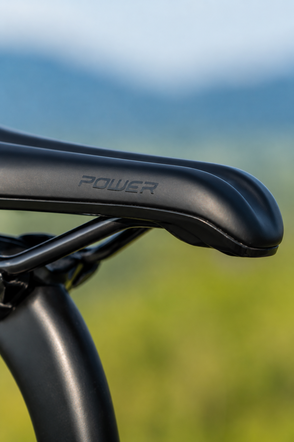 Detalle de un sillín de bicicleta con la palabra 'POWER' grabada en él, en un entorno al aire libre con fondo borroso de cielo y naturaleza.