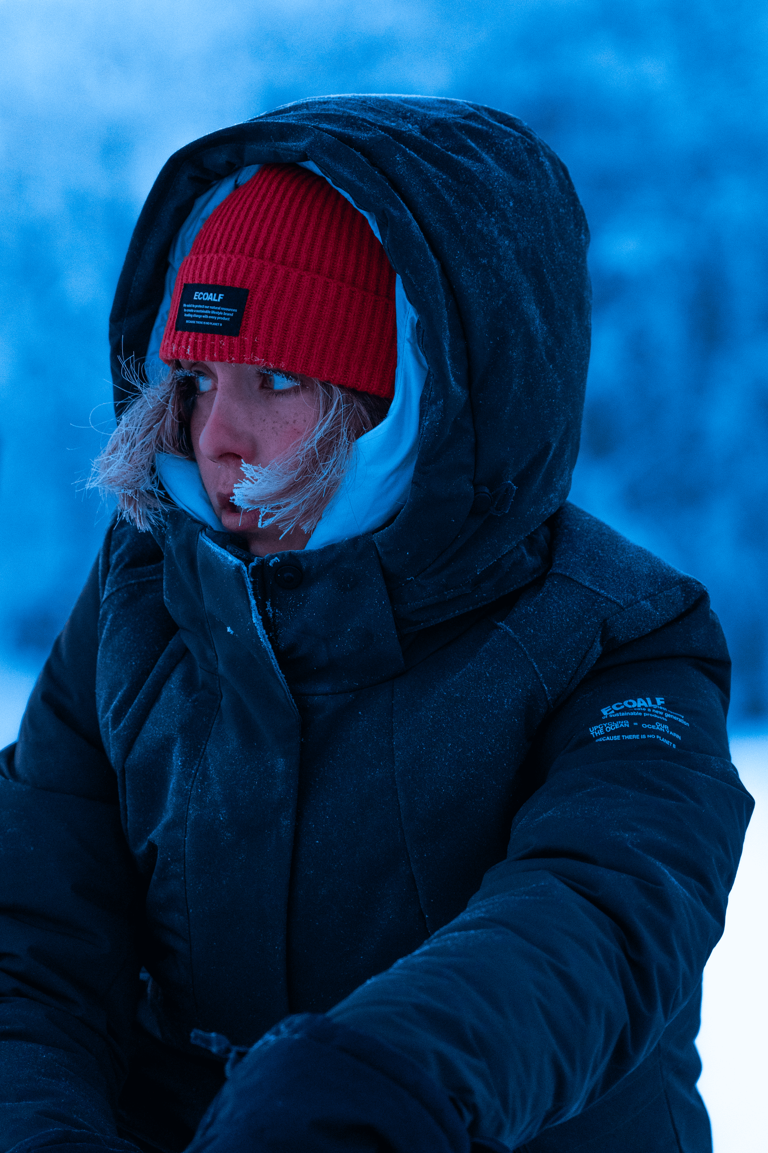 Mujer con ropa de abrigo en un entorno frío, con gorro rojo y chaqueta negra, en un paisaje invernal con fondo azul.