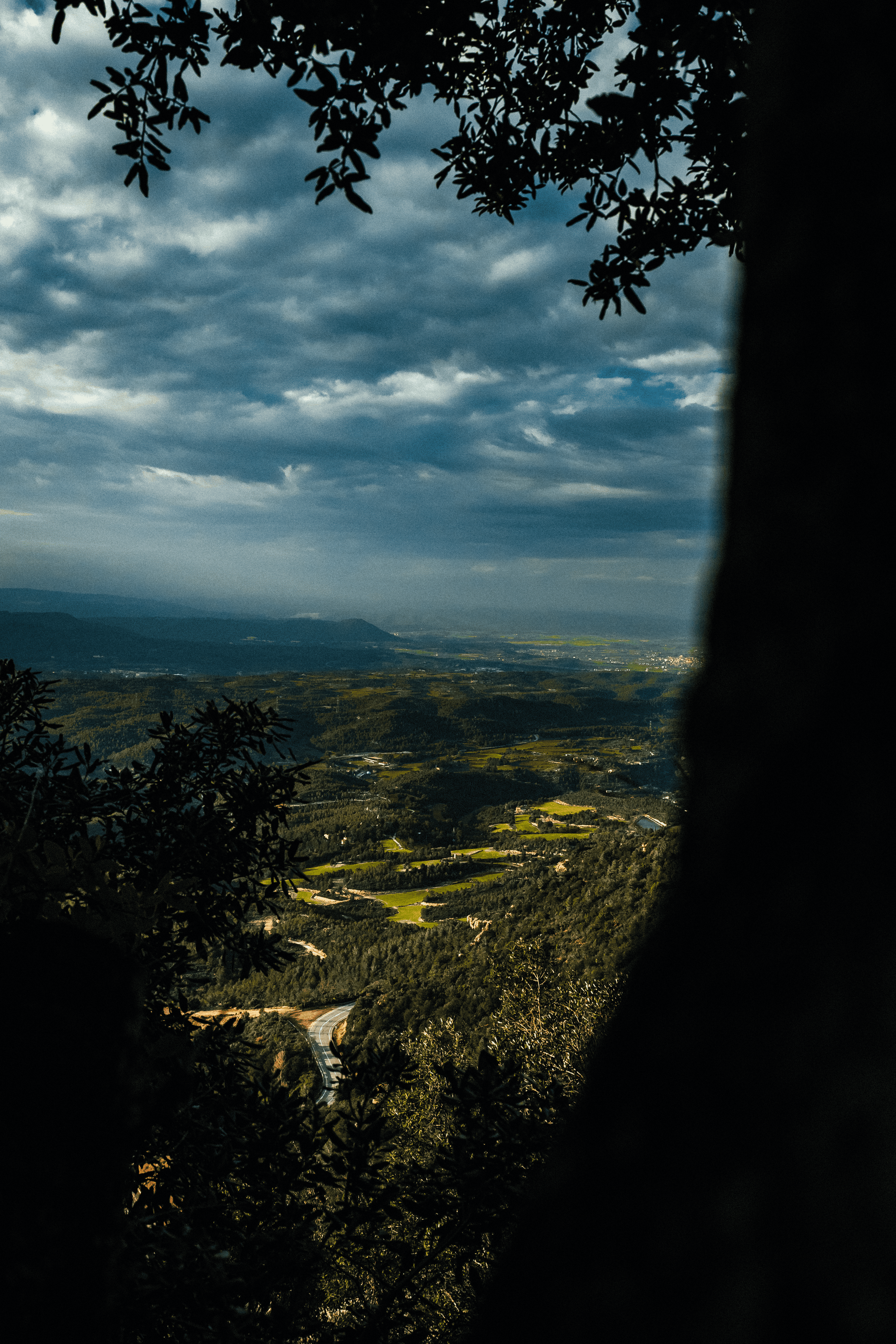 Vista panorámica de montañas y campo vista a través de una abertura rodeada de árboles, cielo nublado.