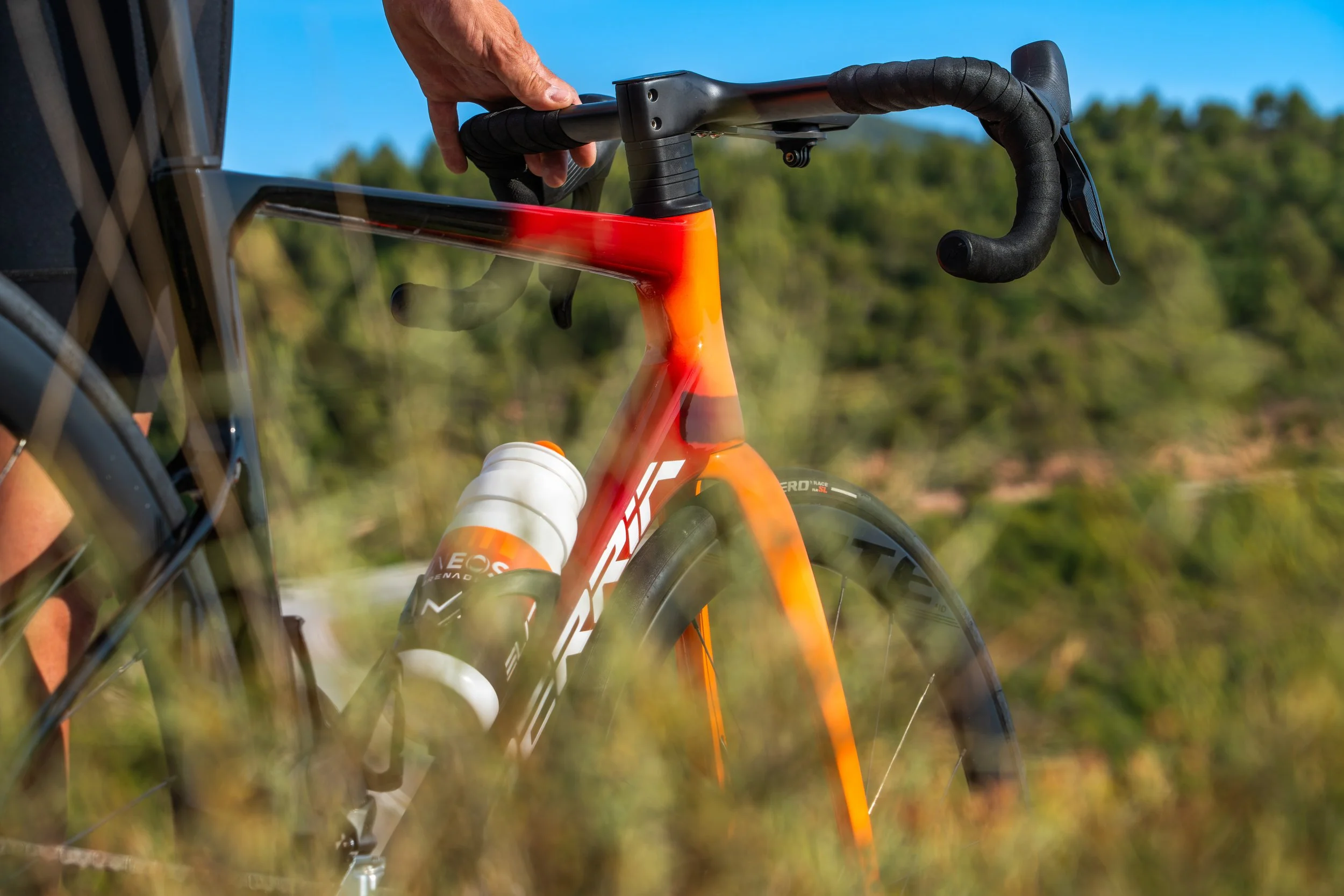 Persona ajustando el manubrio de una bicicleta de color naranja en un entorno al aire libre con cielo azul y árboles en el fondo.