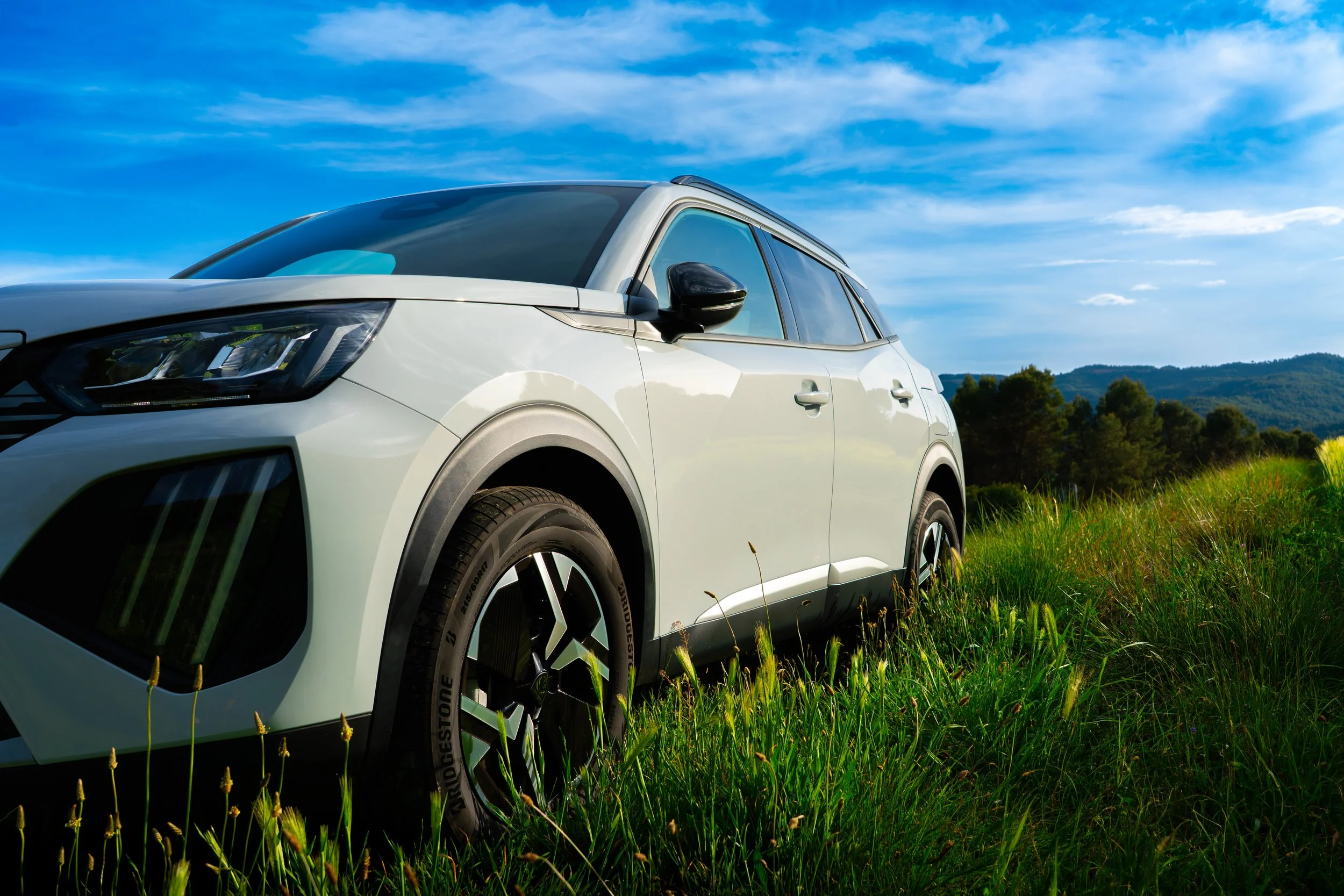 Coche blanco en un campo con colinas y árboles, cielo despejado con algunas nubes.