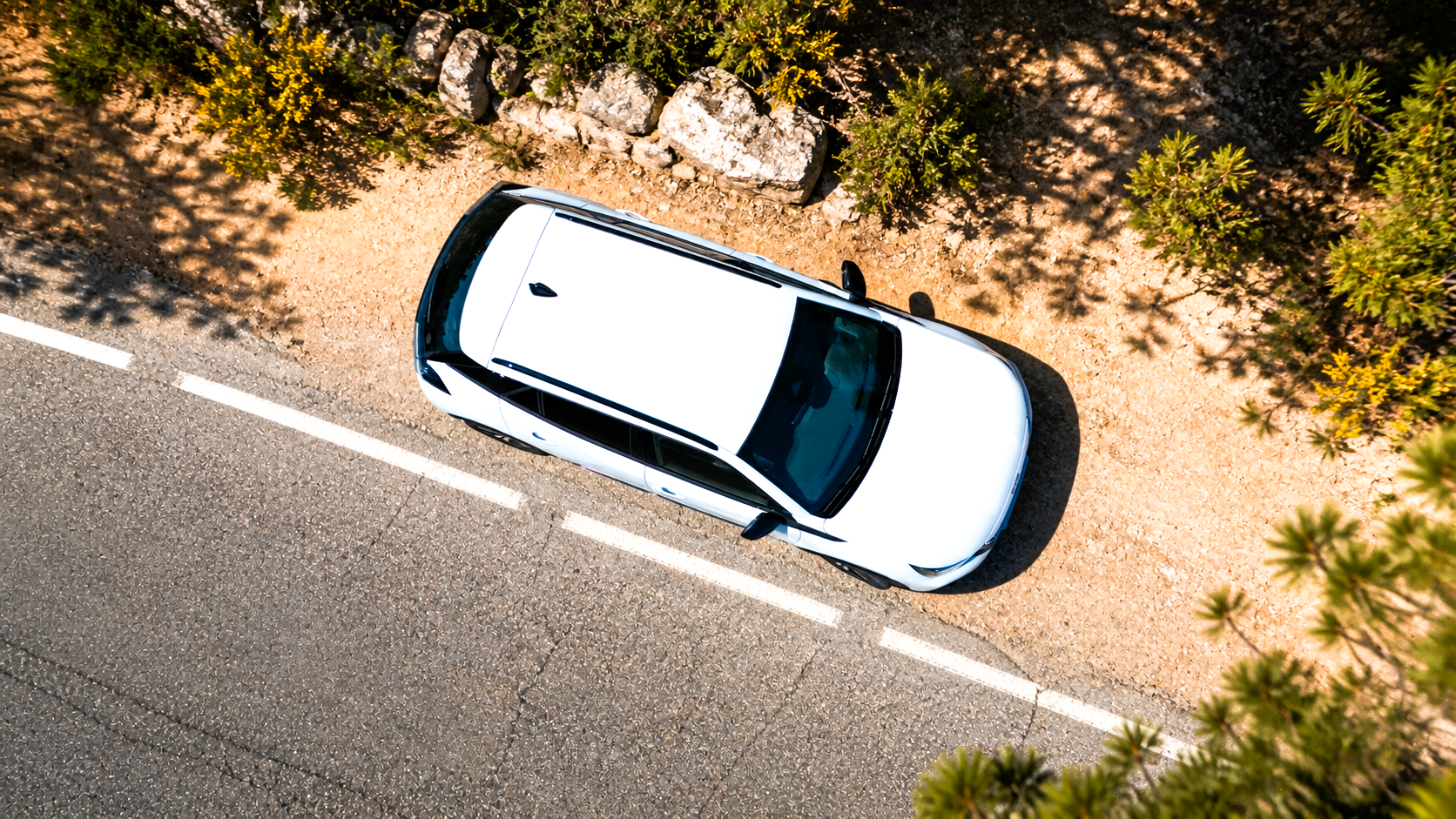 Vista aérea de un coche blanco estacionado a un lado de una carretera, rodeado de arbustos y rocas.