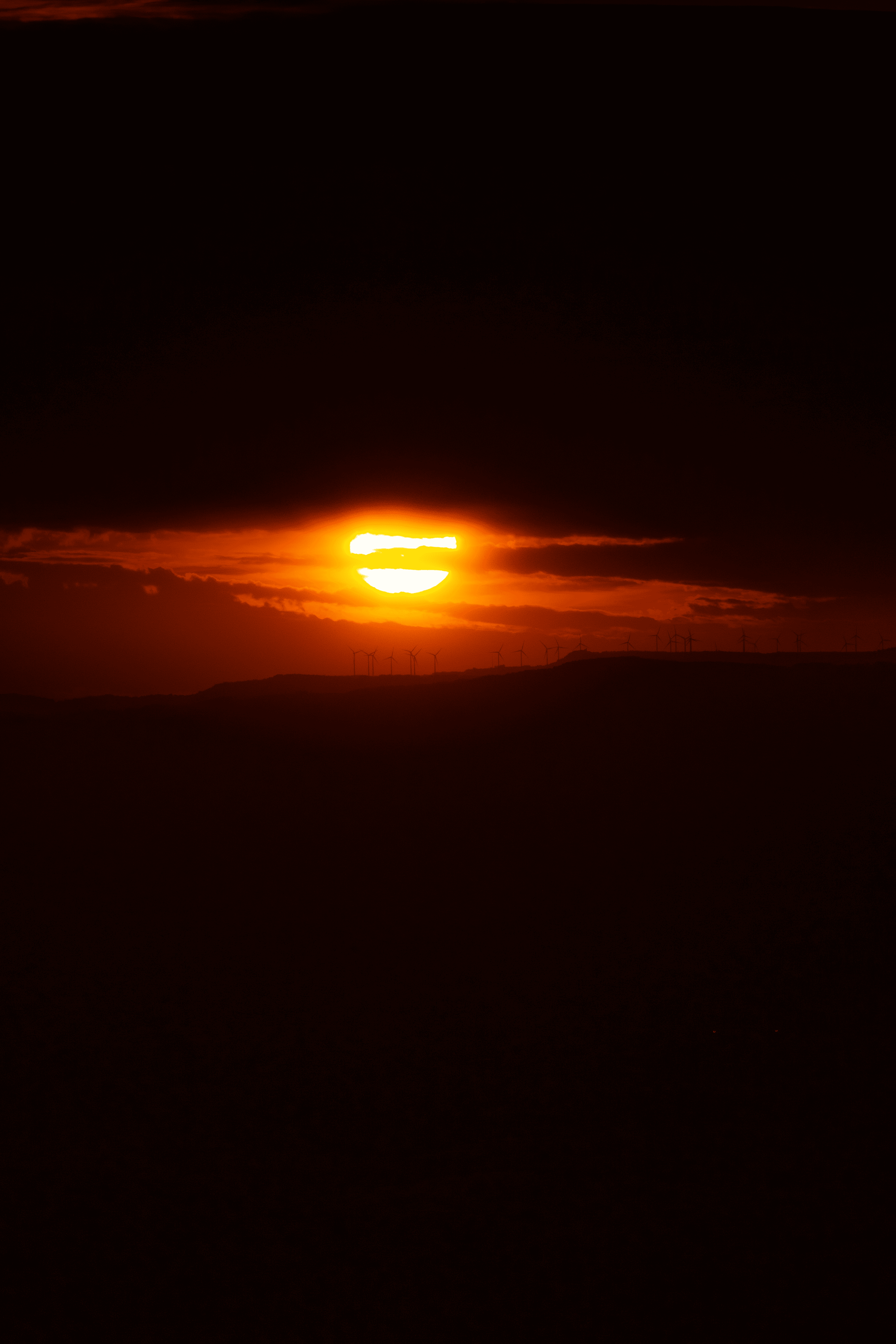 Atardecer con sol parcialmente cubierto por nubes y aerogeneradores en el horizonte.