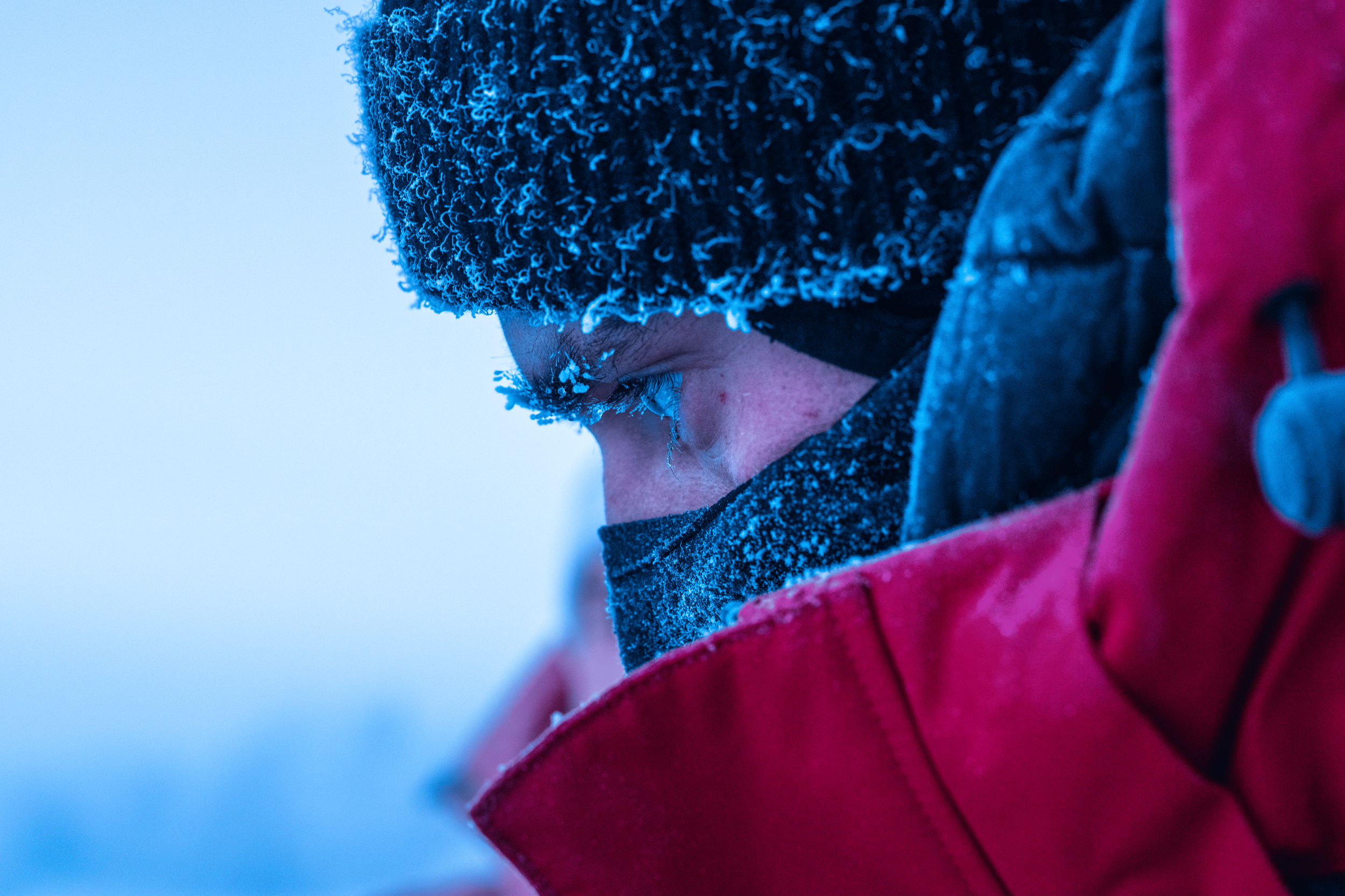 Persona con gorro, mascarilla y chaqueta roja en un entorno frío con escarcha en su rostro y ropa.