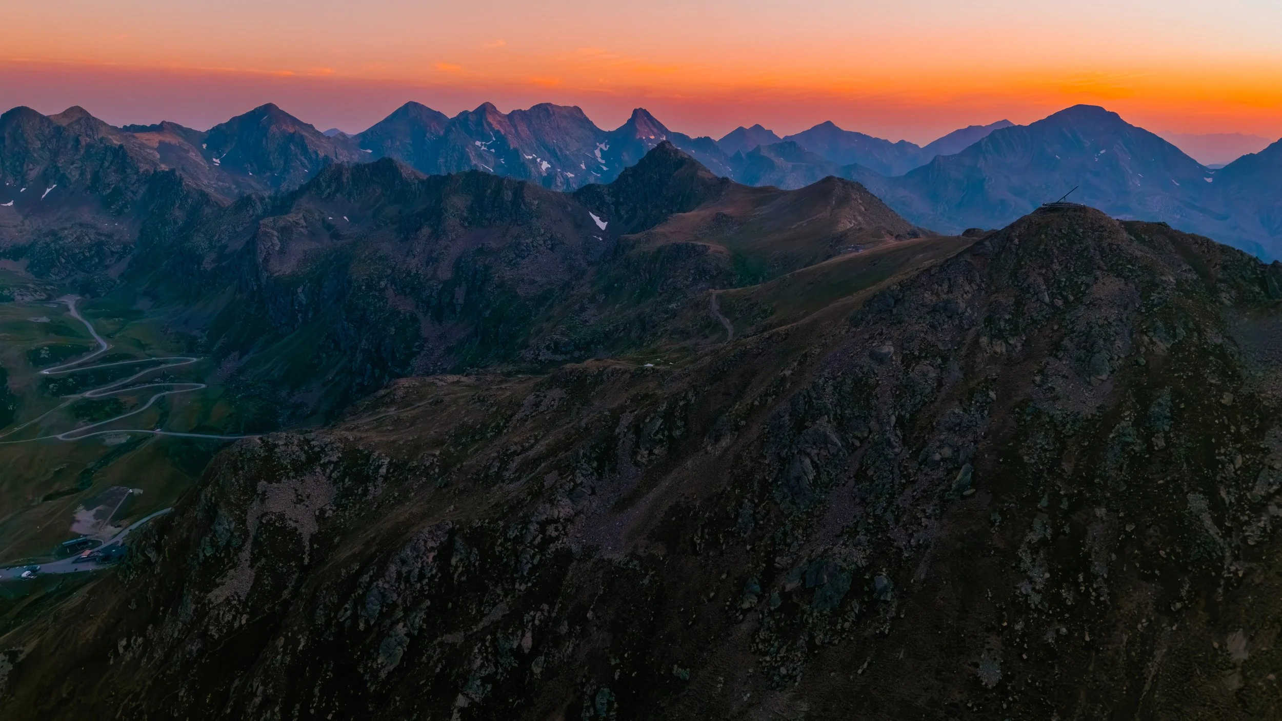 Vista panorámica de montañas en el atardecer con cielos anaranjados y caminos serpentinos en el valle.