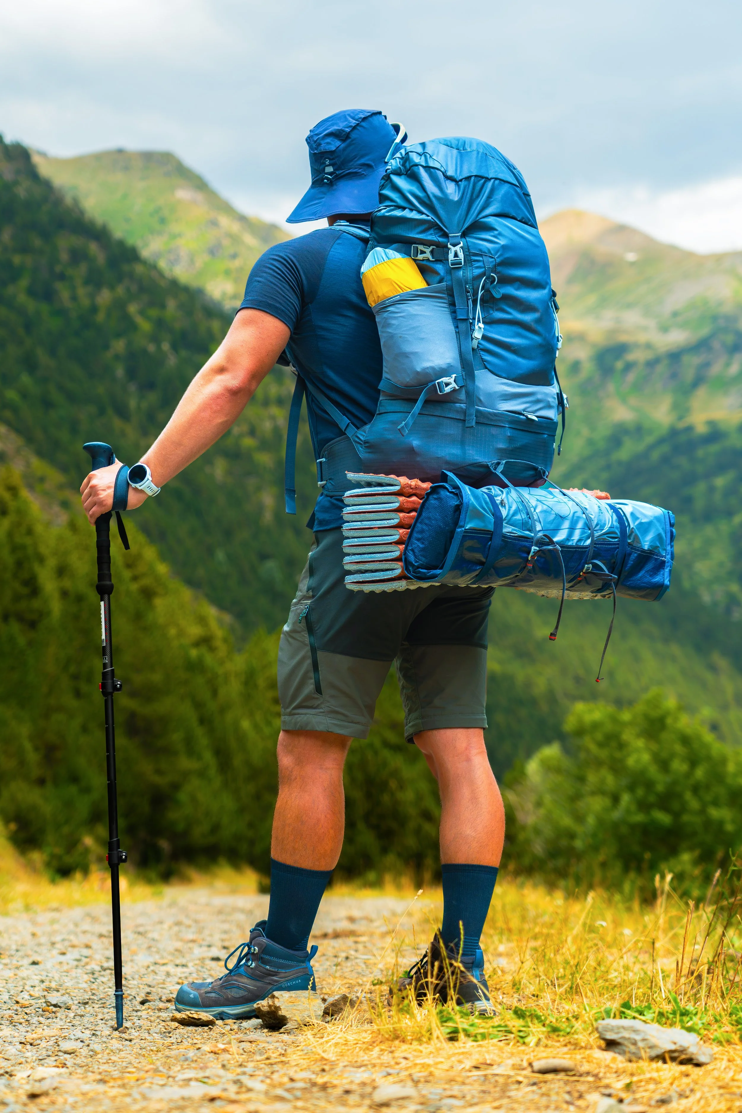 A hiker con mochila grande y gorro azul, usando bastón y ropa deportiva, caminando por un sendero en un paisaje montañoso con vegetación y cielo nublado.