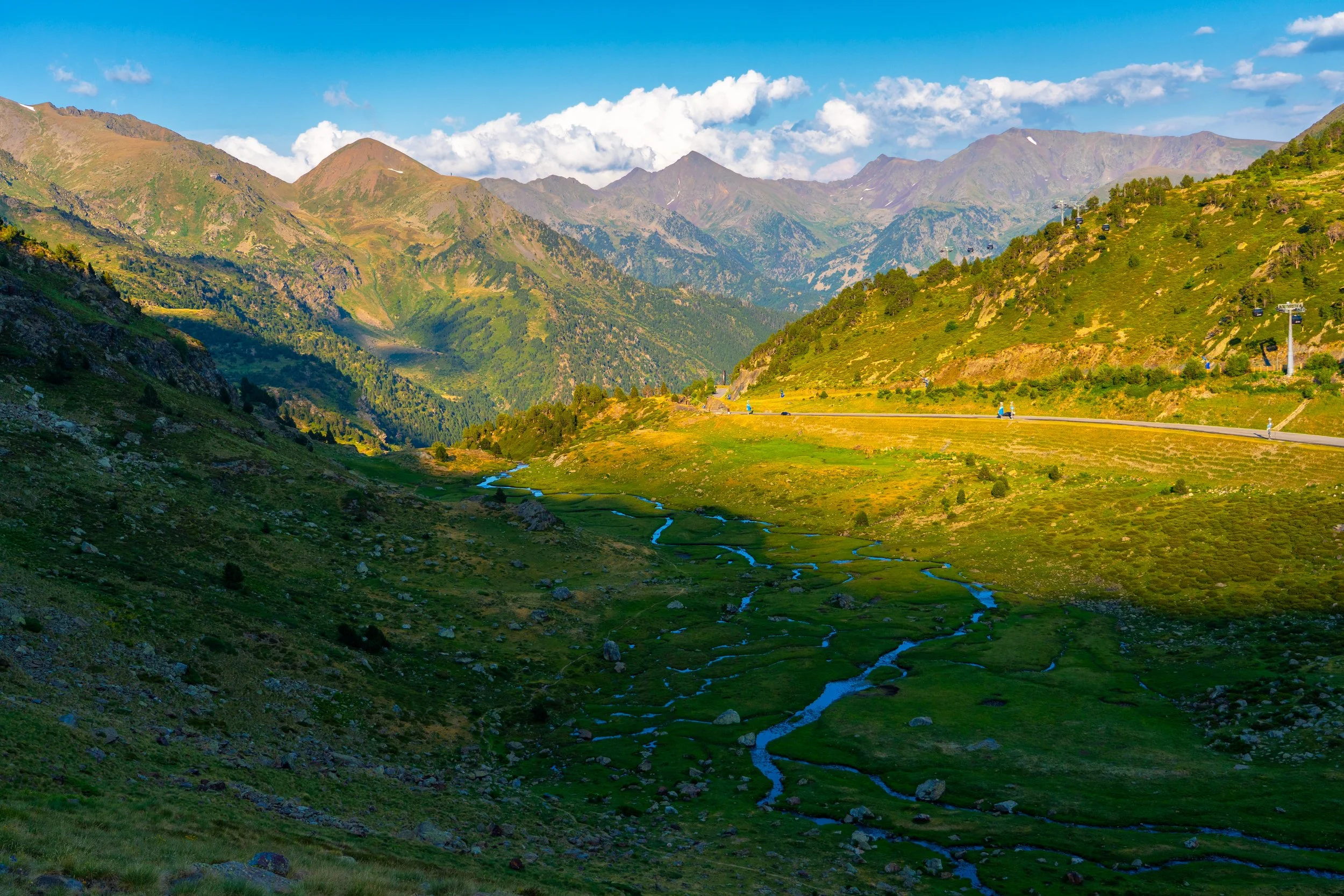 Paisaje montañoso con picos altos, vegetación verde en las colinas y un río que serpentea por el valle, bajo un cielo azul con algunas nubes.