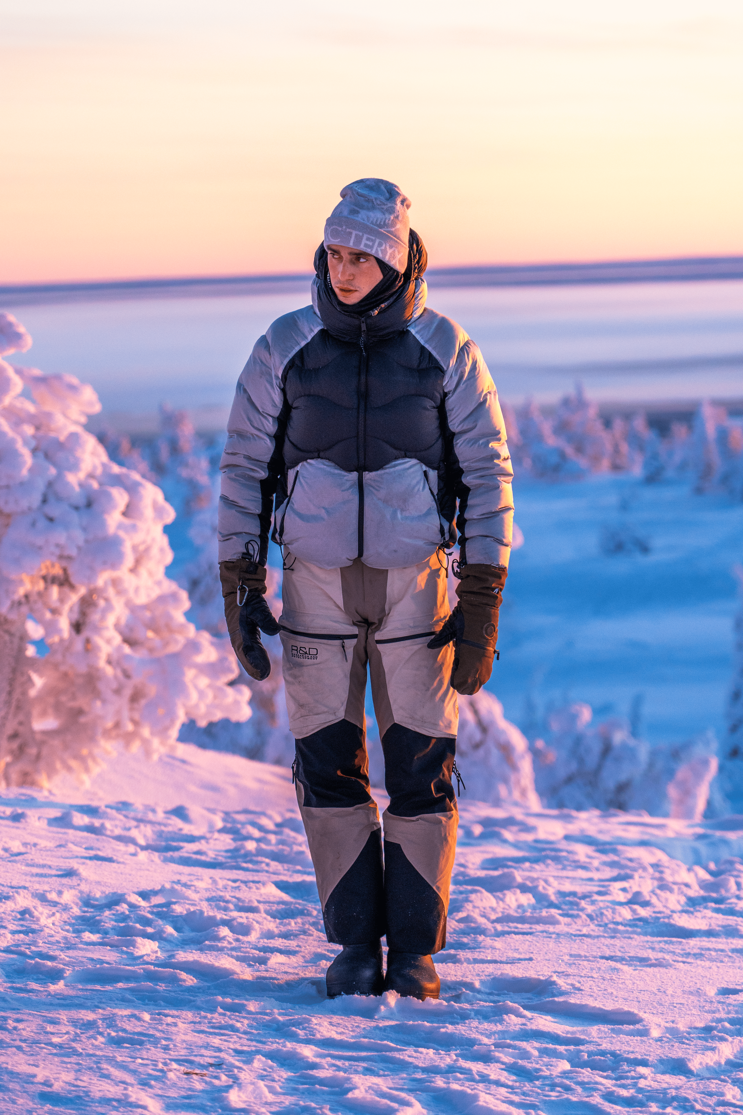 Persona caminando en un paisaje nevado durante el atardecer, vestida con ropa de invierno negra y gris, en un entorno con árboles cubiertos de nieve.