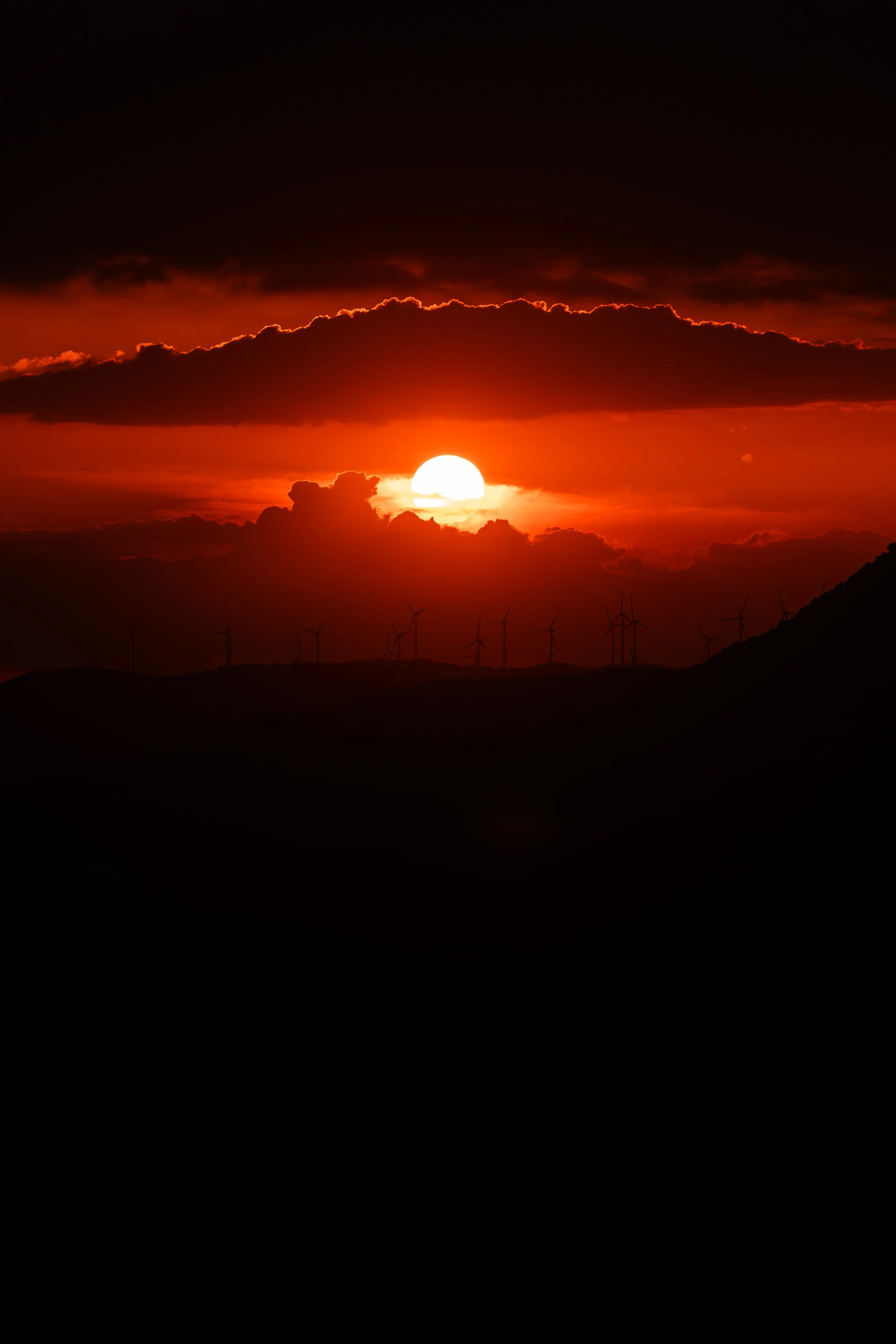 Atardecer con el sol en el horizonte, nubes oscuras y molinos de viento en las colinas.