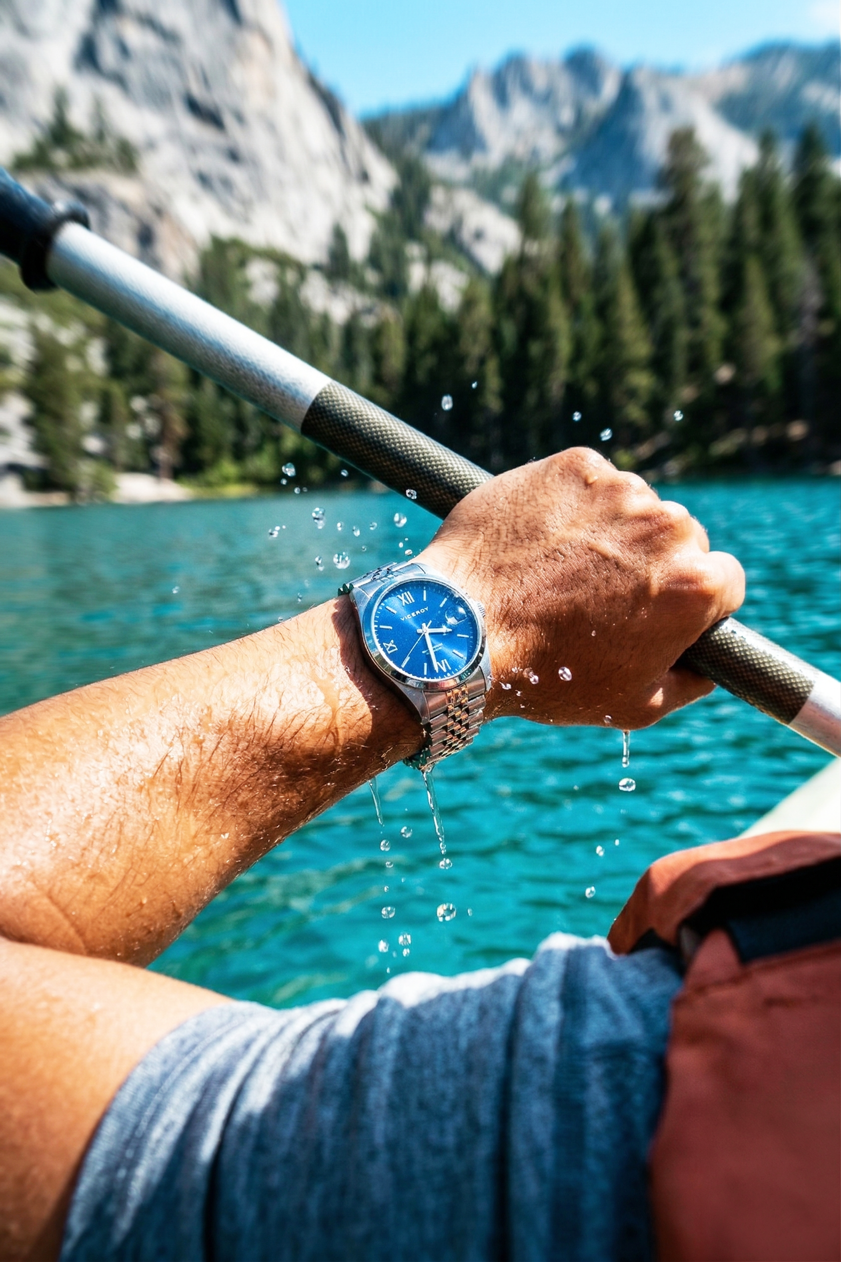Mano sujetando un remo en un lago con montañas y árboles de fondo, lleva un reloj de pulsera de correa metálica.