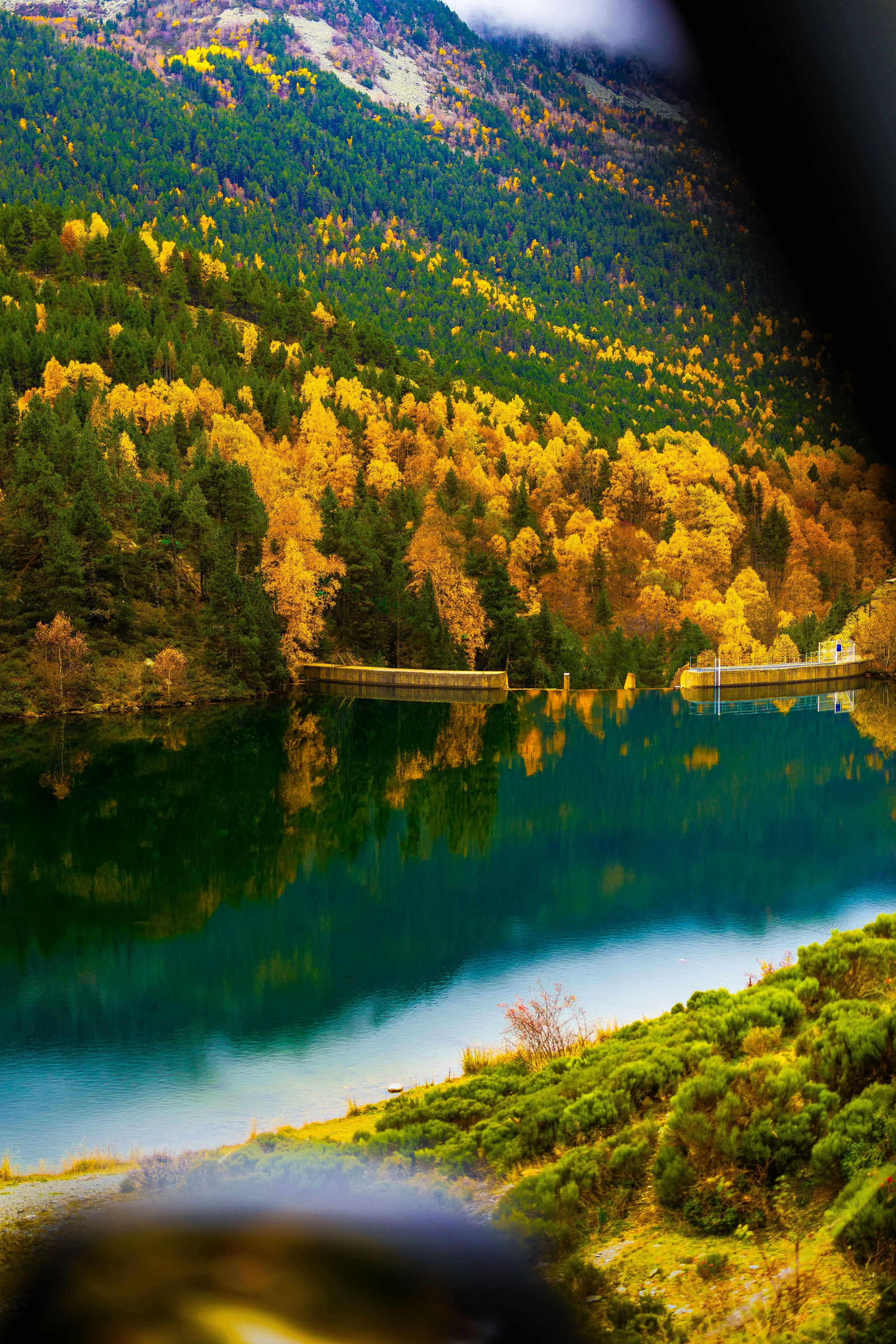 Paisaje de montañas con árboles en colores otoñales y reflejo en un lago, rodeado de vegetación y nubes en el cielo.