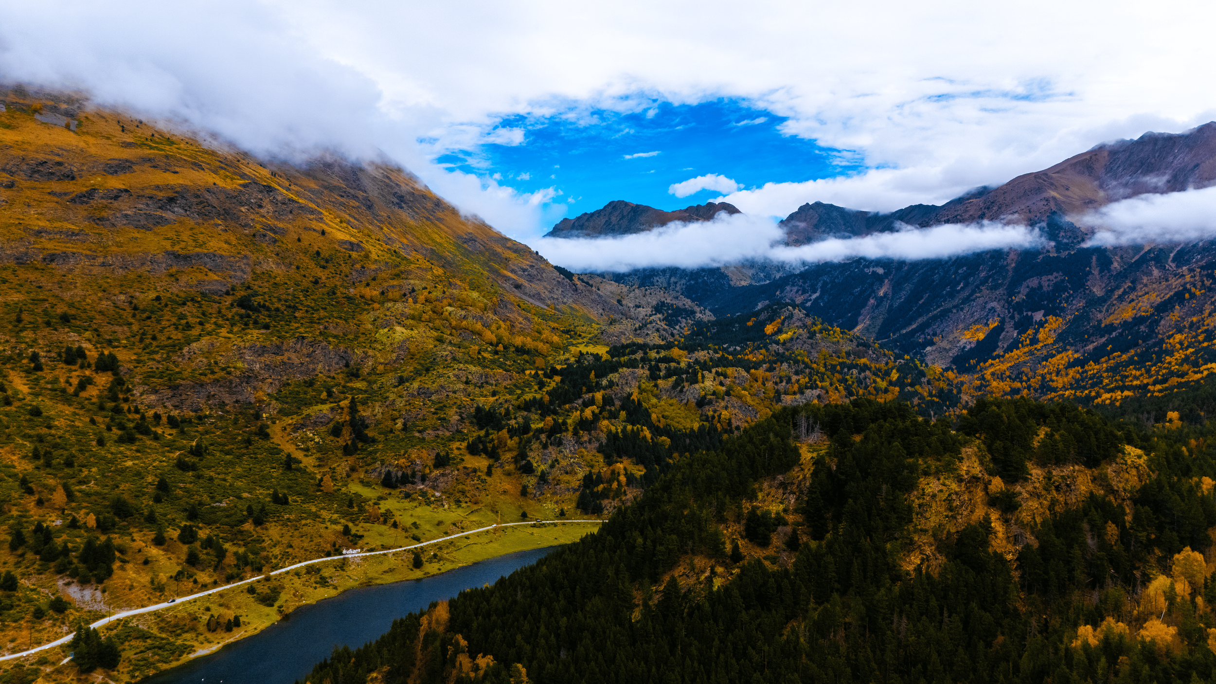 Montañas cubiertas de vegetación con nubes y cielo azul, un lago en primer plano y una carretera que lo atraviesa