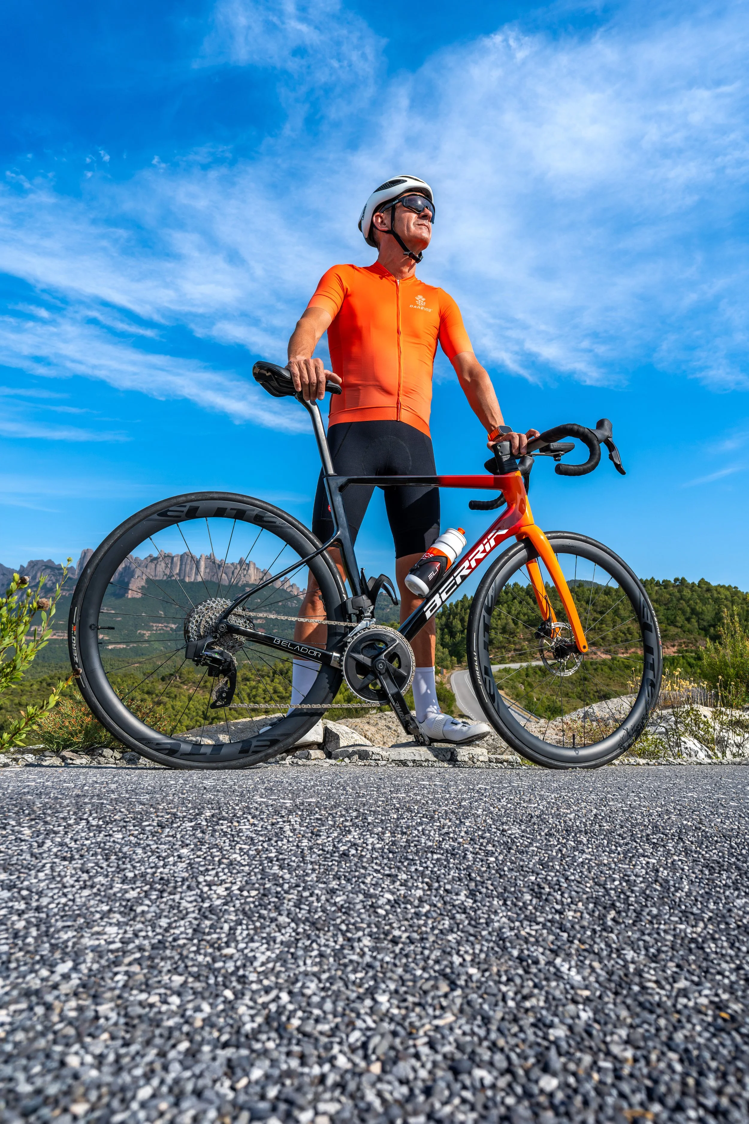 Hombre con ropa deportiva y casco de ciclismo descansando en un entorno montañoso con cielo despejado, sosteniendo su bicicleta de ruta con paisaje de montañas en el fondo.