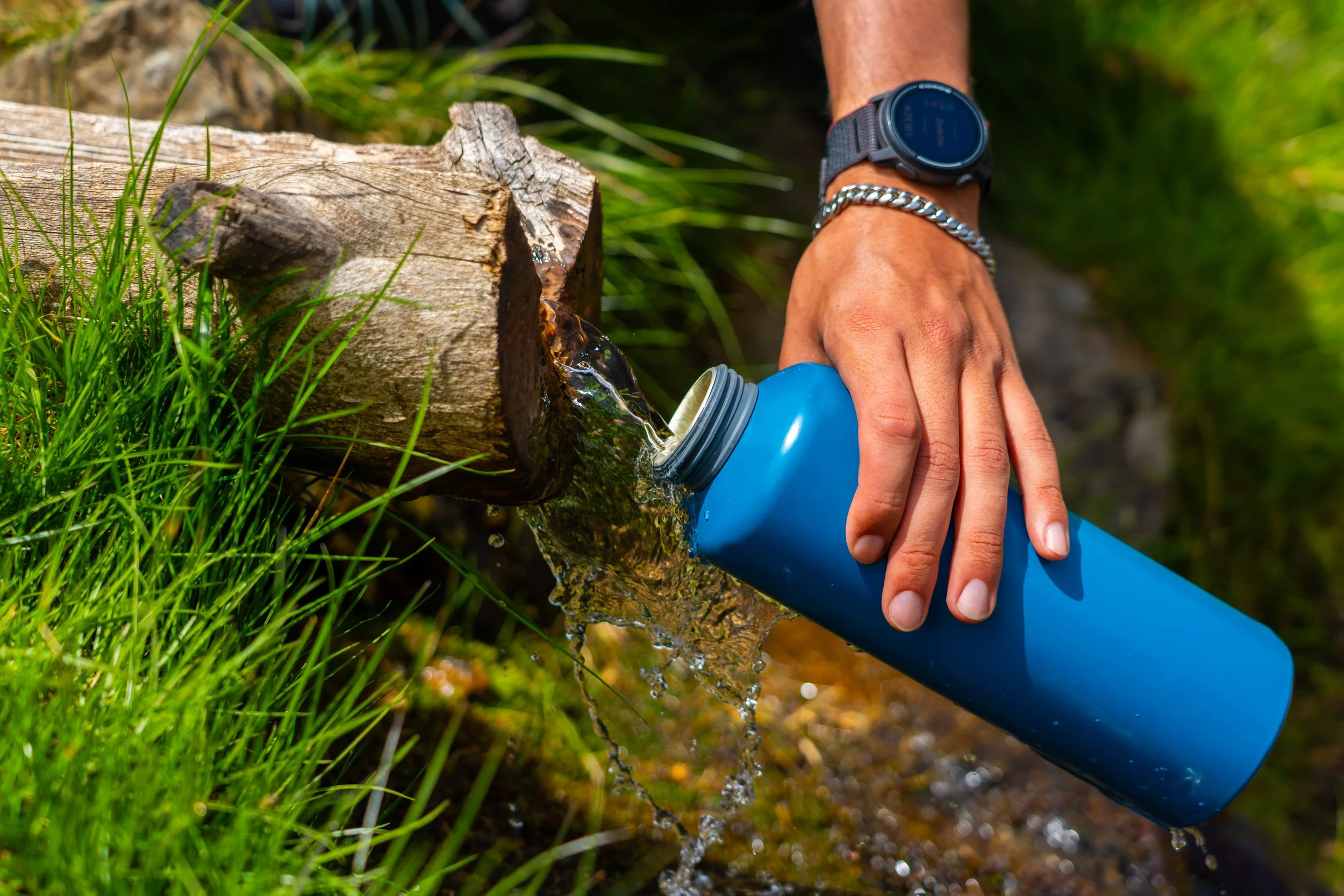 Una mano está llenando una botella azul con agua de una tubería en un entorno natural con hierba y árboles.