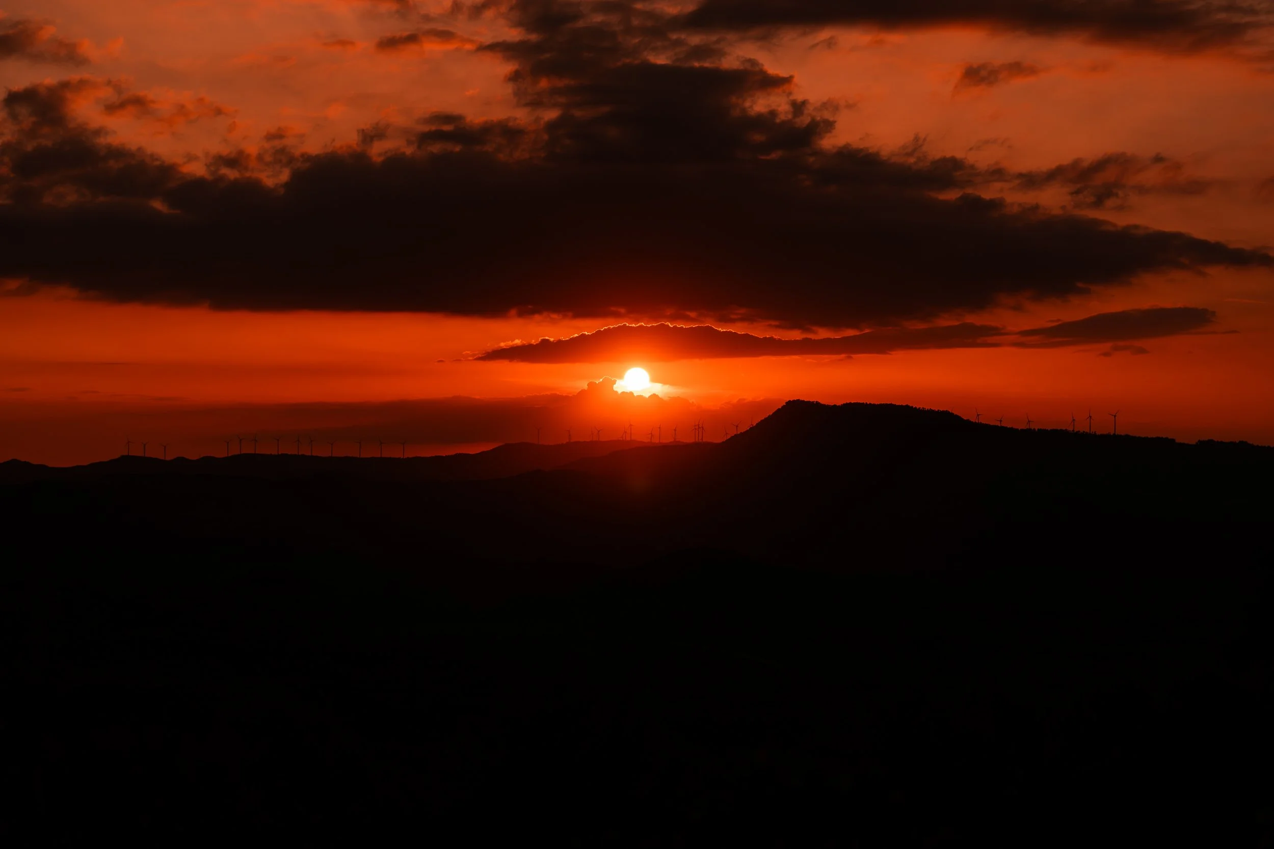 Atardecer con el sol ocultándose detrás de las nubes y un paisaje con molinos de viento en primer plano.