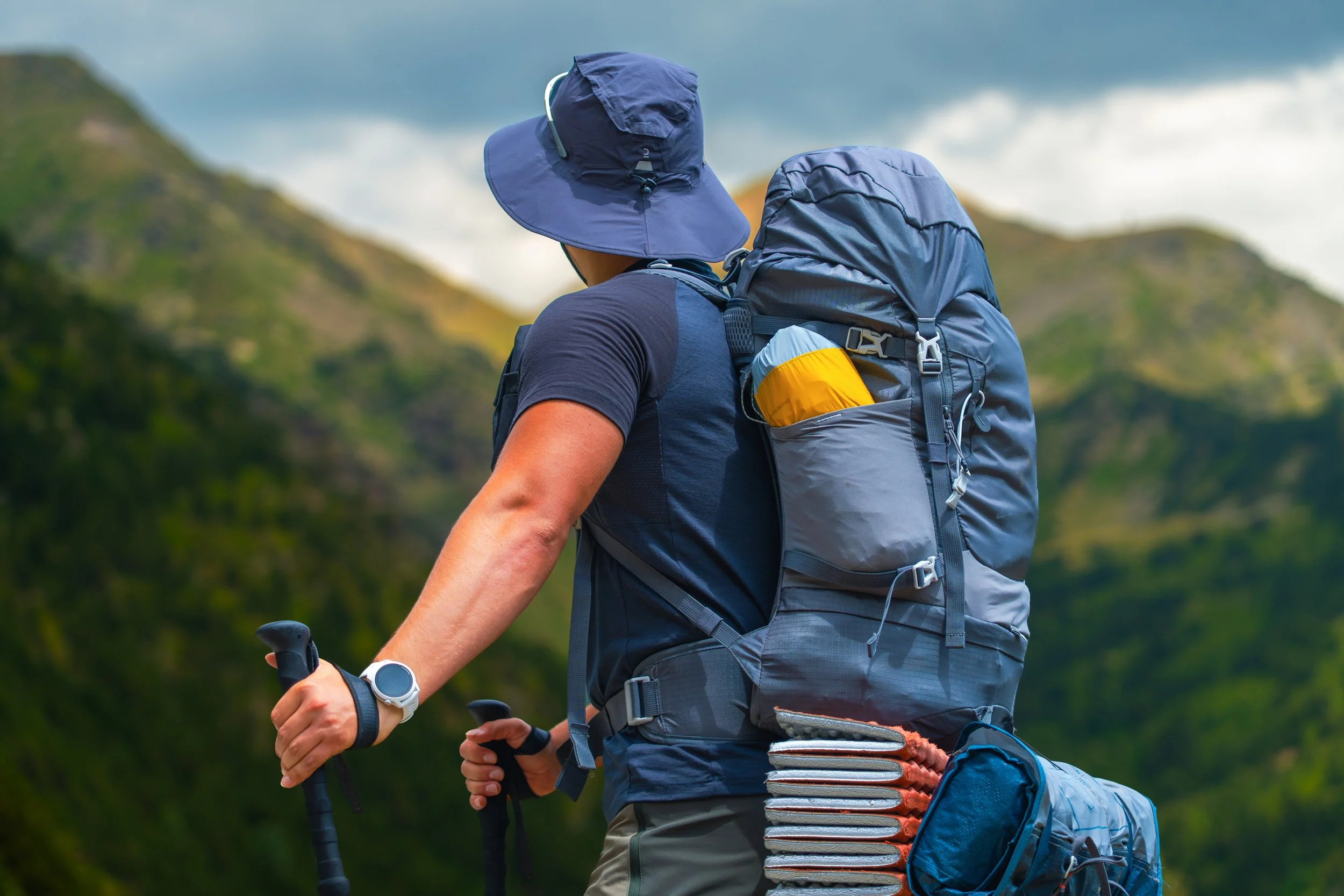 Persona con sombrero y mochila de senderismo, caminando por un área natural montañosa.
