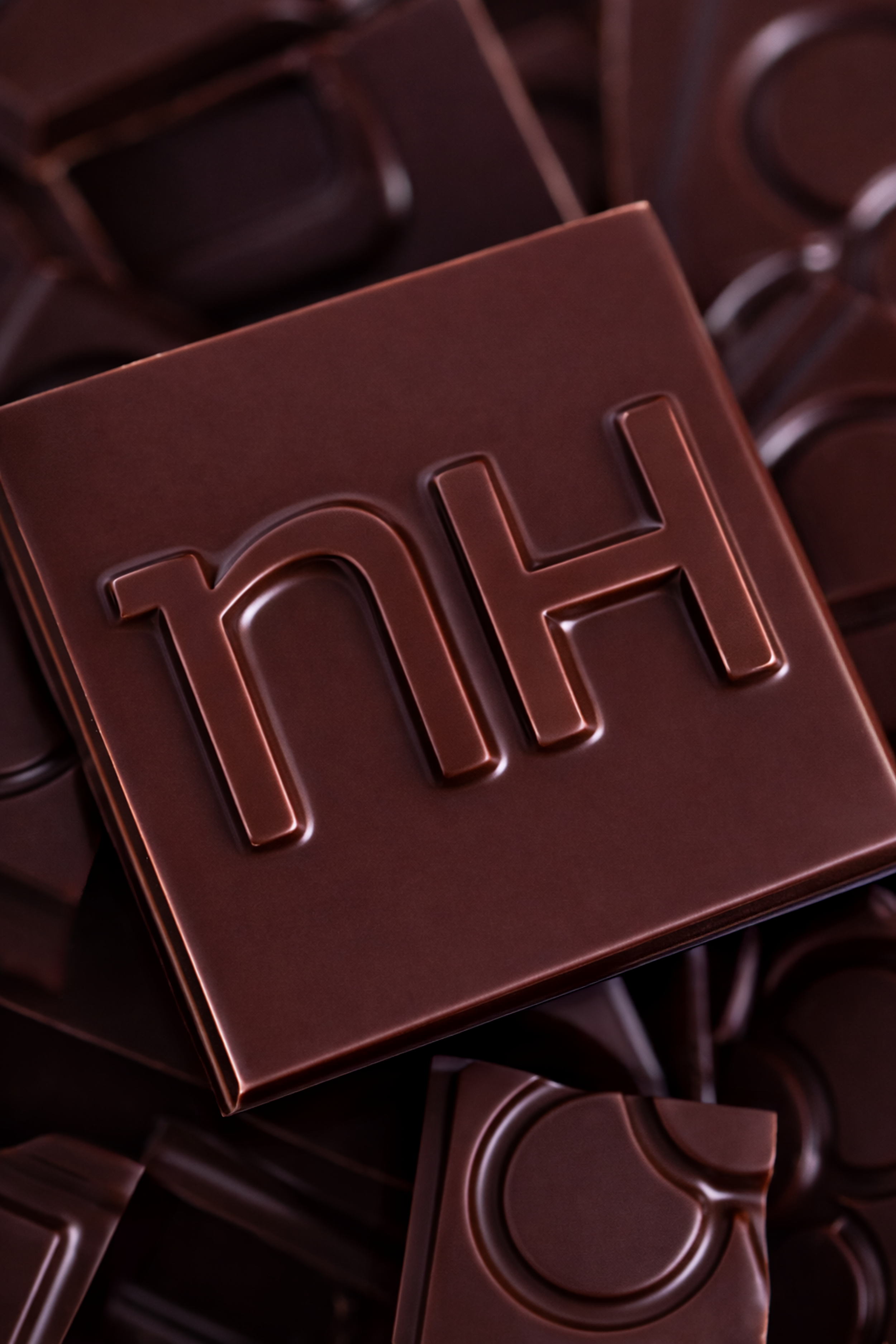 Caja de chocolate con la marca "nh" en relieve en la parte superior, rodeada de otras piezas de chocolate.