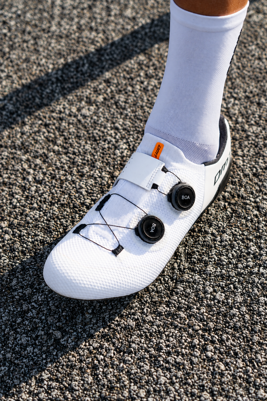 Un zapato de ciclismo blanco con calas y pedalieres en una superficie de asfalto, con un calcetín blanco visible.
