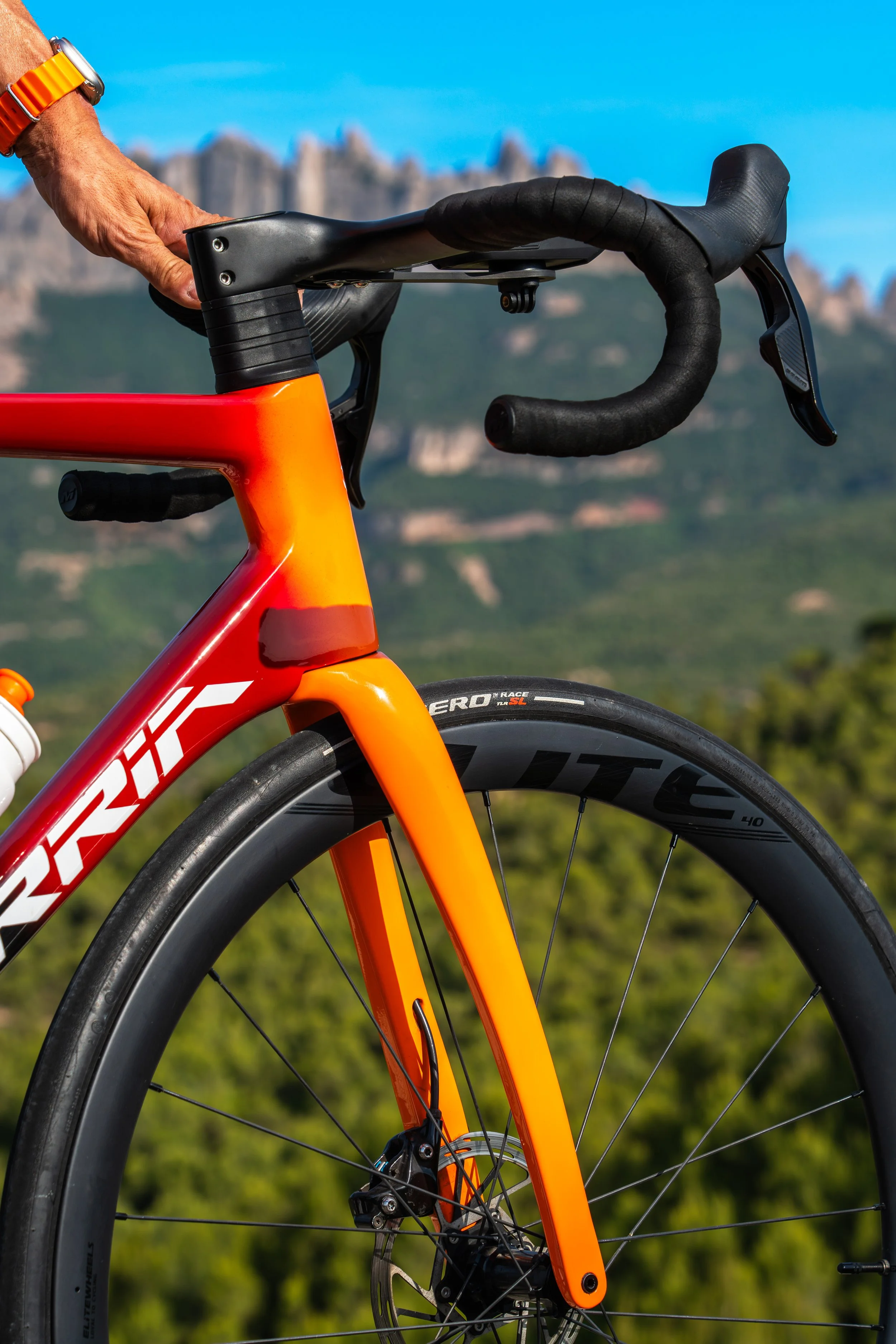 Primera parte de una bicicleta de carreras con cuadro naranja y componentes de fibra de carbono, en un paisaje con montañas y cielo azul al fondo.
