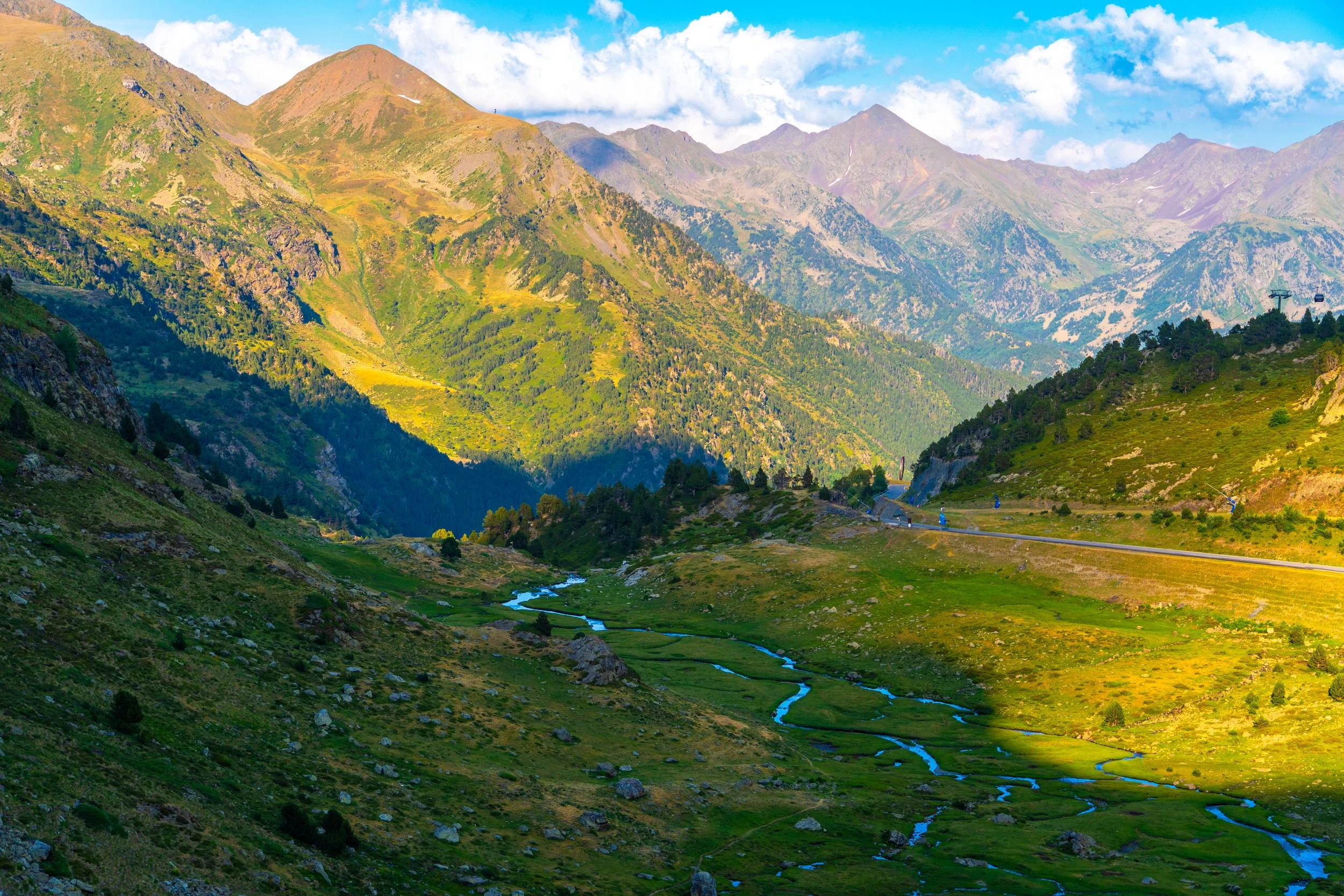 Paisaje montañoso con picos elevados y vegetación verde, un río que serpentea por el valle y un cielo parcialmente nublado.
