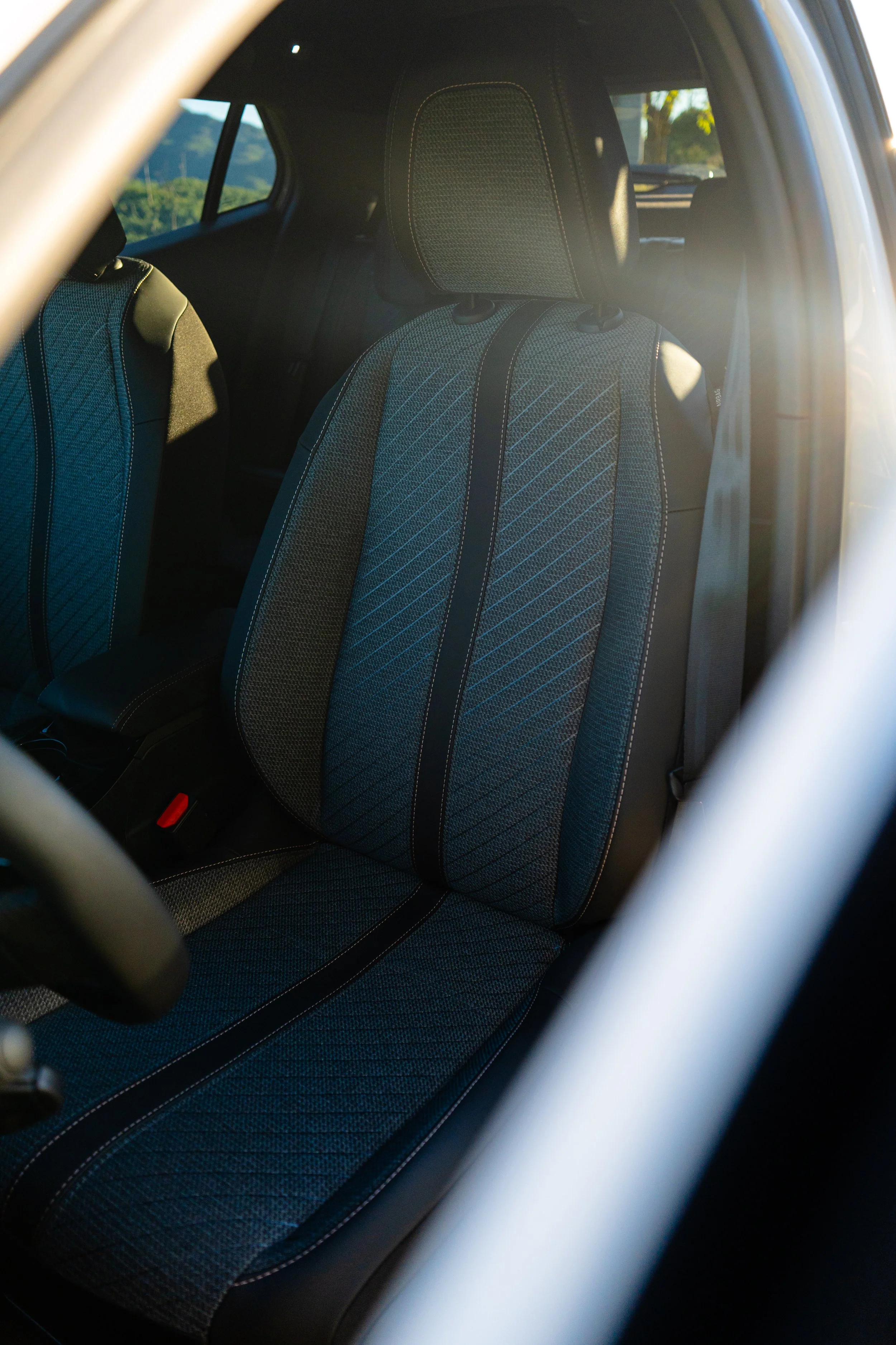 Asiento de coche con tapizado en tela de color gris con patrones y costuras blancas, visto a través de la ventana del vehículo al atardecer.