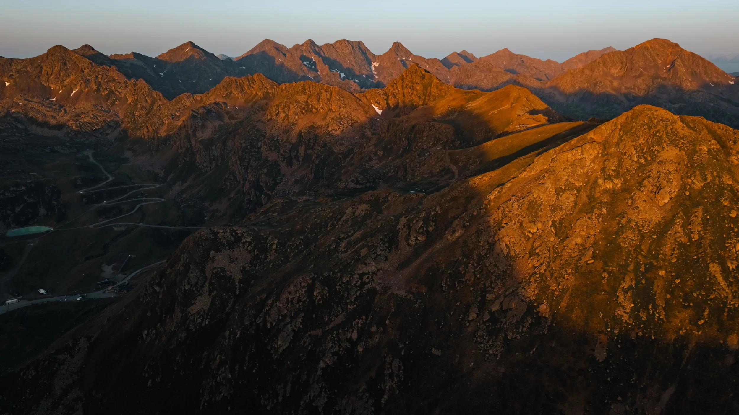 Vista panorámica de cadenas montañosas de forma irregular con picos altos y pendientes empinadas, iluminadas por la luz dorada del atardecer, con algunos picos con restos de nieve y senderos en las laderas.