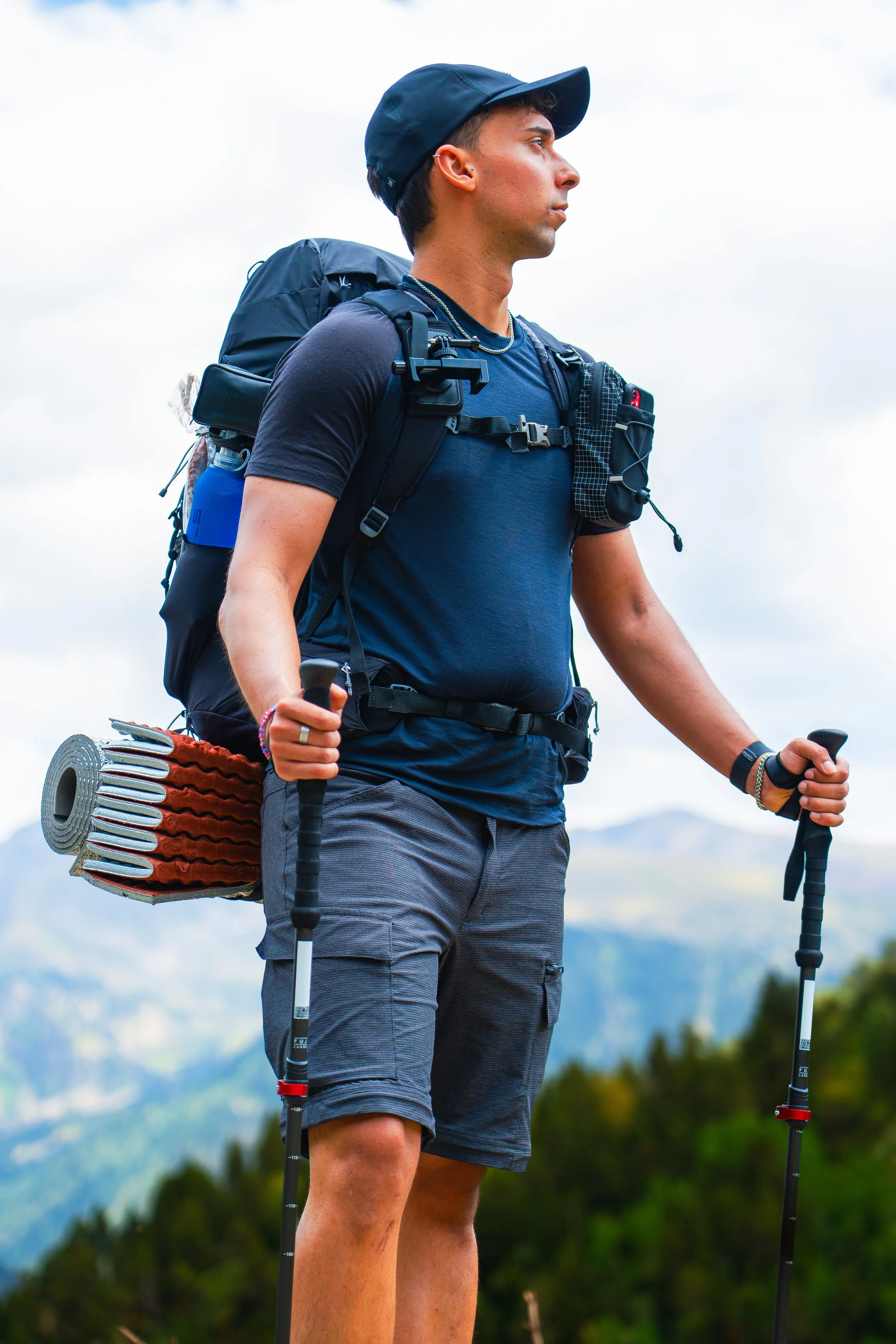 Un hombre joven de pie en un paisaje montañoso con collarines, mochila grande y bastones de senderismo, mirando hacia un lado con fondo de nubes y naturaleza.