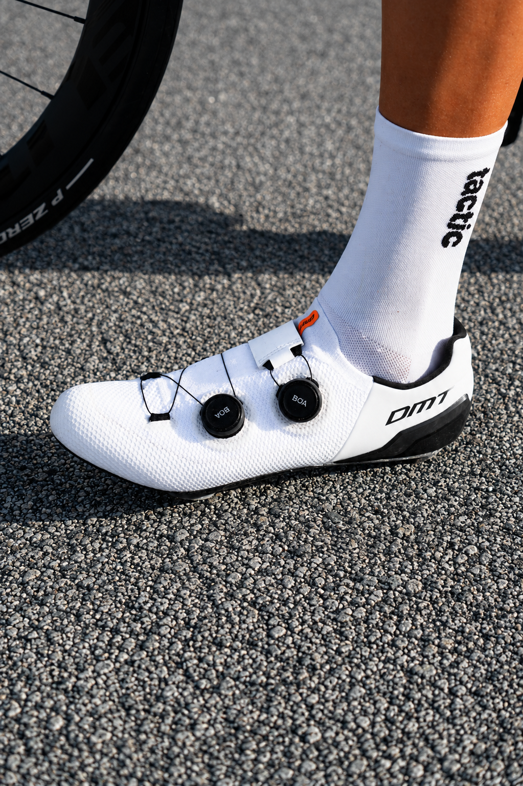 Zapatos de ciclismo blancos con calzado técnico y un ciclista con calcetas blancas que tienen la palabra 'tactic' escrita. La rueda de una bicicleta está parcialmente visible en la esquina superior izquierda. El suelo es de asfalto.