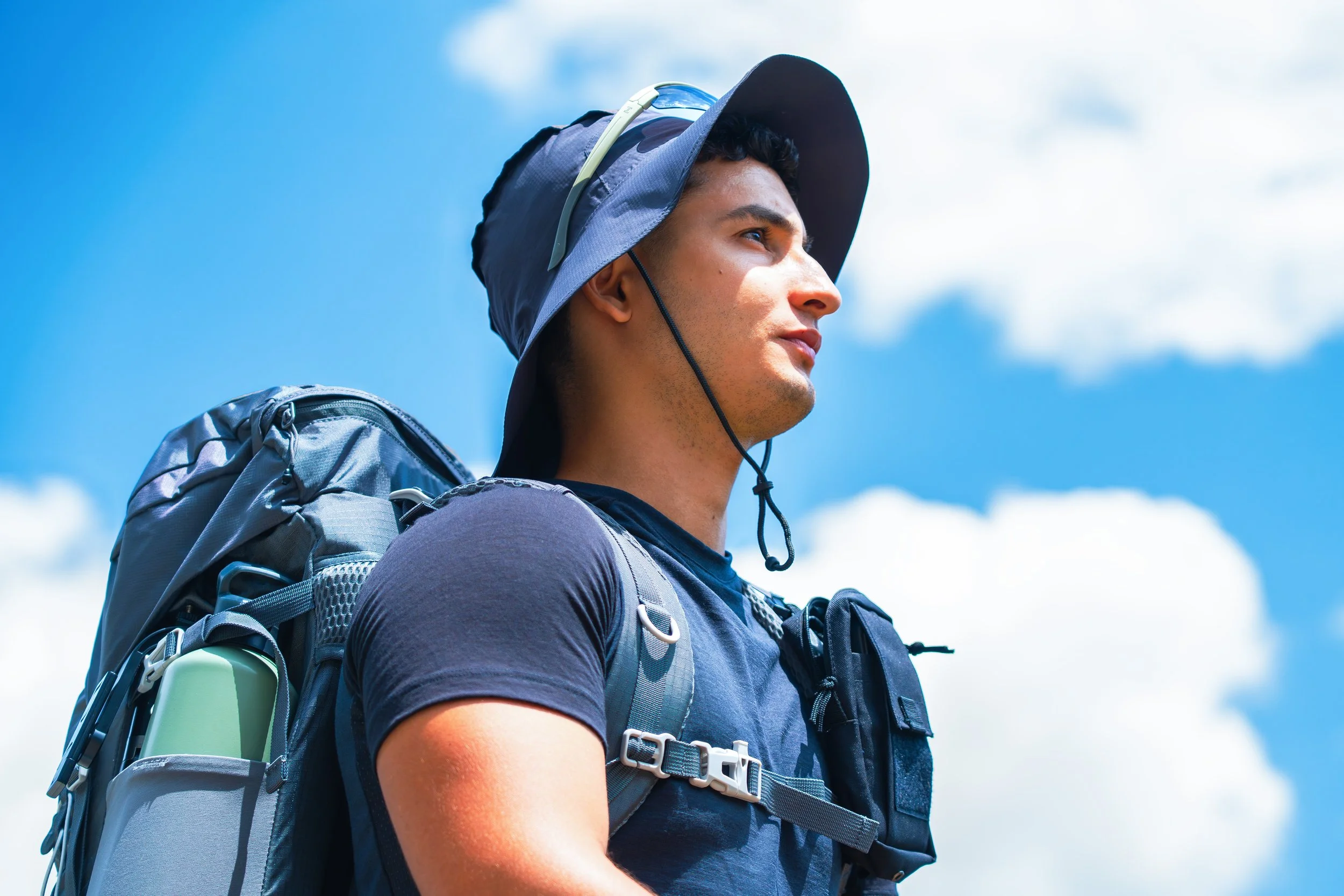 Joven hombre con mochila y gorra de explorador observando el cielo azul con nubes