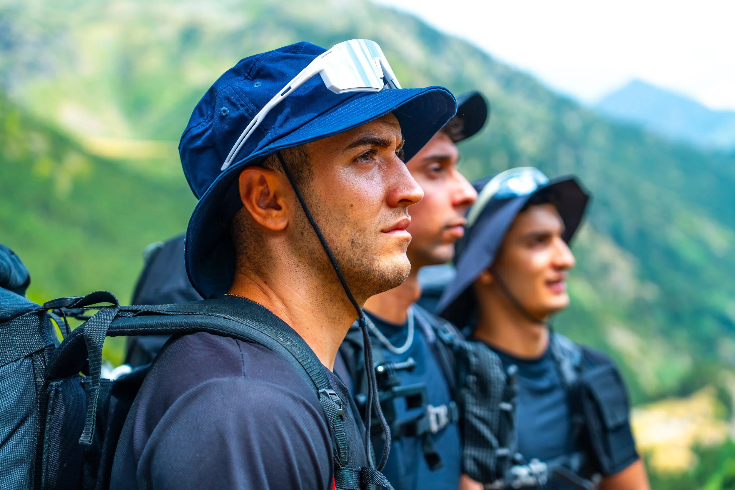 Grupo de personas jóvenes con mochilas y gorros, de perfil, en naturaleza, en fila, mirando hacia adelante.