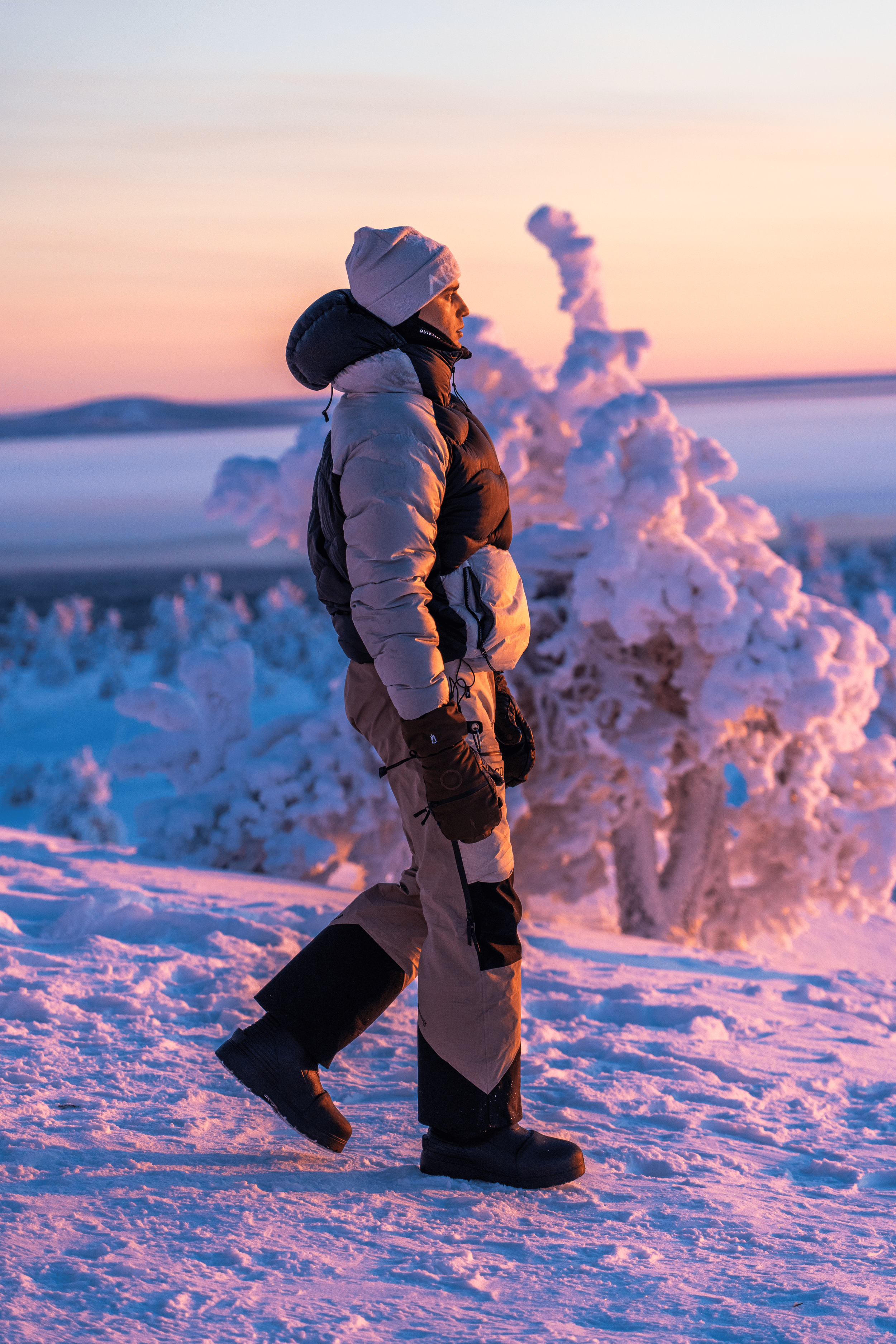 Persona haciendo senderismo en un paisaje nevado al atardecer, con árboles cubiertos de nieve y un cielo con tonos rosas y naranjas.