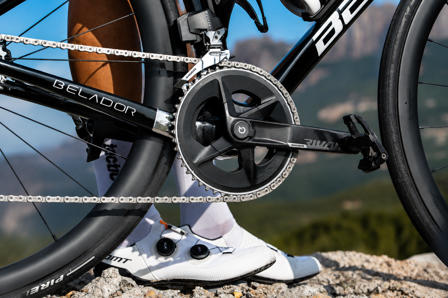 Ciclista con zapatos blancos y pedales automáticos, bicicleta de ruta con marco negro y componentes de fibra de carbono, en un terreno de piedras con fondo montañoso.
