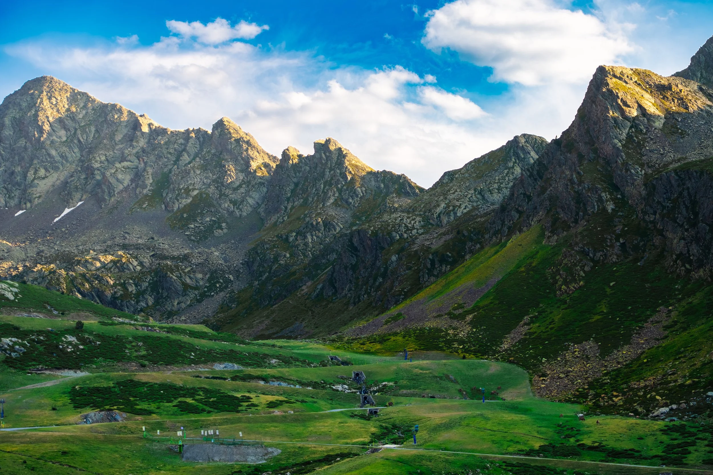 Montañas altas con picos rocosos bajo un cielo azul con nubes dispersas y un valle verde en primer plano.
