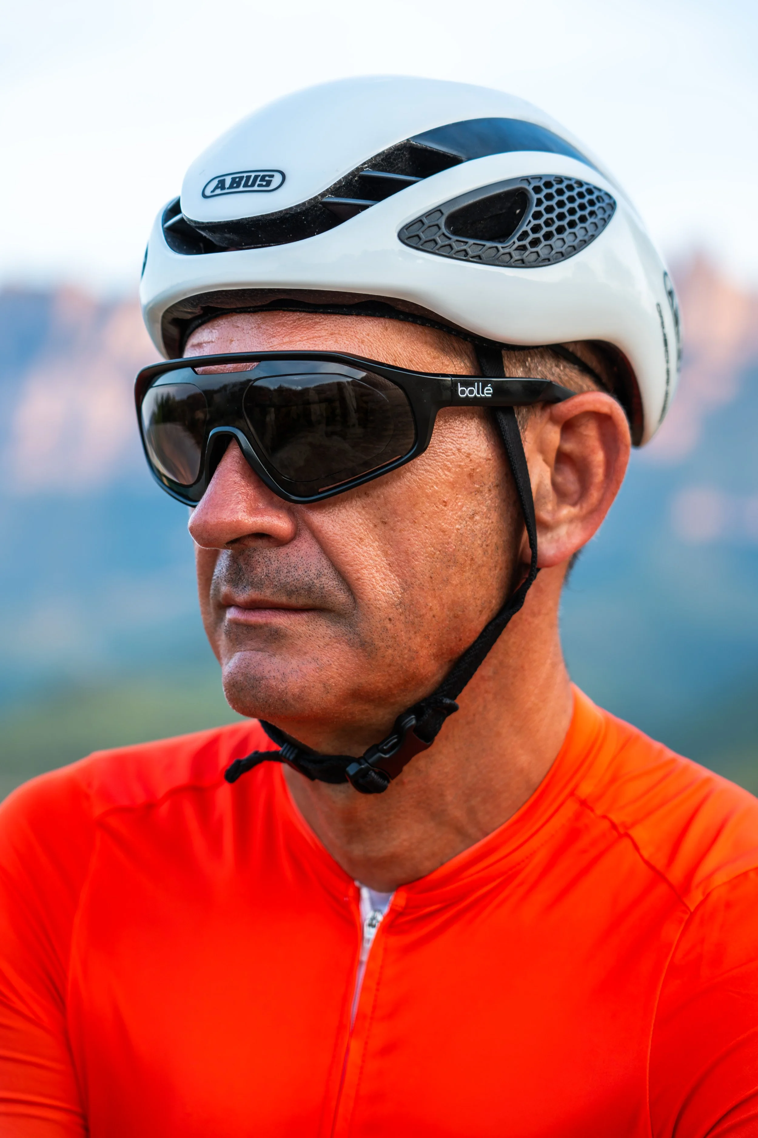 Hombre con gafas deportivas y casco de ciclismo de color blanco, vistiendo una camiseta naranja, en un entorno exterior