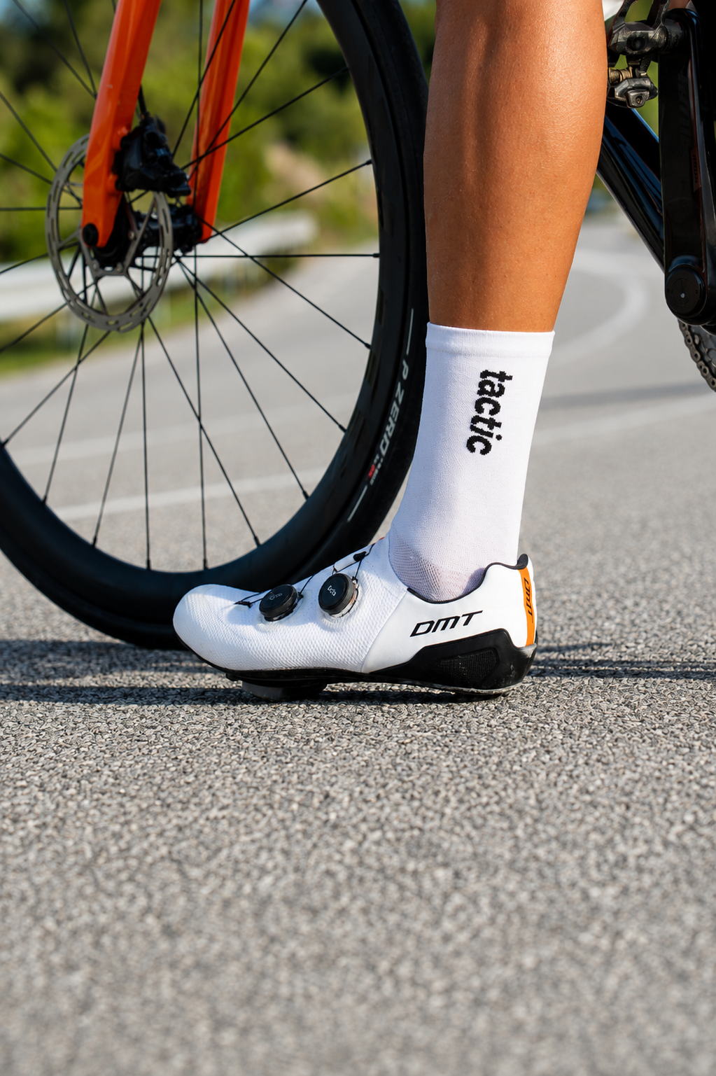 Pie del ciclista con calceta blanca que tiene la palabra 'tactic' en negro, calzado de ciclista blanco con LOGO 'DMT' y zapatillas de ciclismo, y parte de la rueda delantera de una bicicleta de ruta con marco naranja.