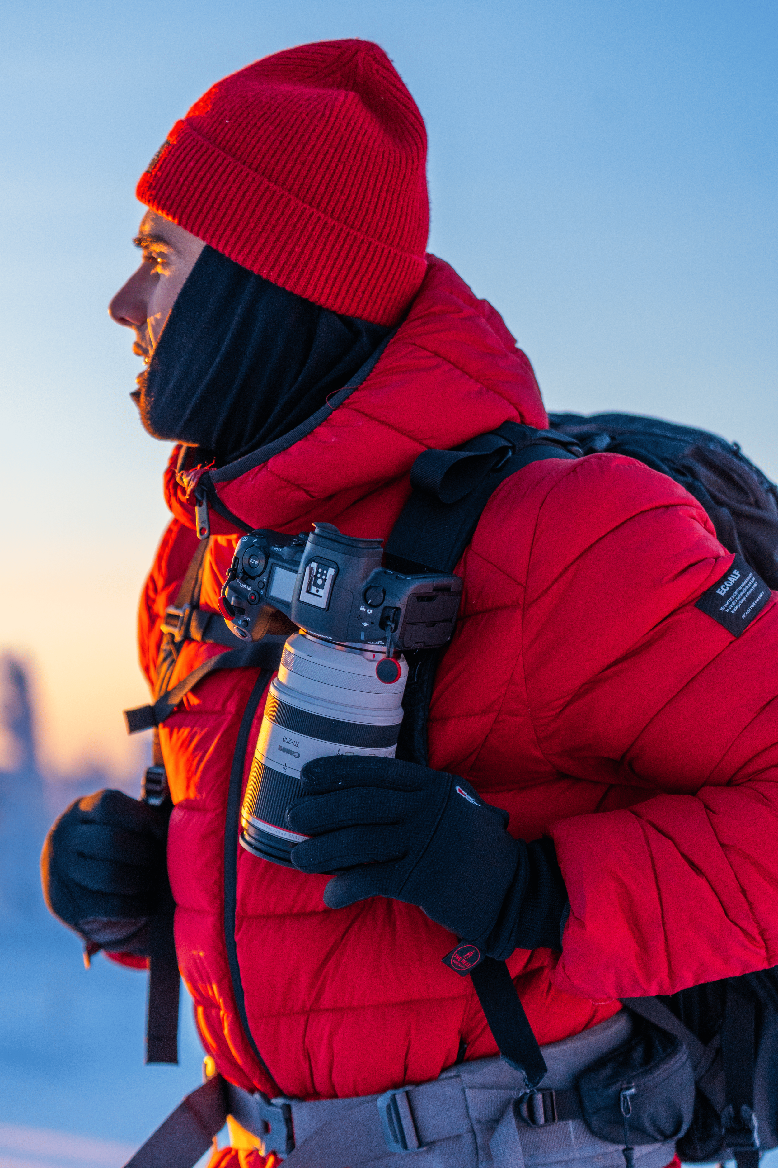 Hombre con ropa de abrigo, gorro y guantes, cargando una cámara con lente, en un paisaje con cielo azul y nieve al fondo.
