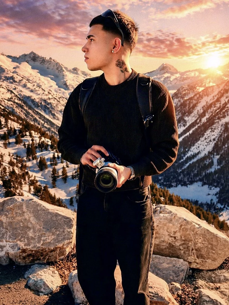 Joven con gafas de sol, camiseta negra y mochila, sosteniendo una cámara, en un entorno montañoso con nieve y árboles, al atardecer.
