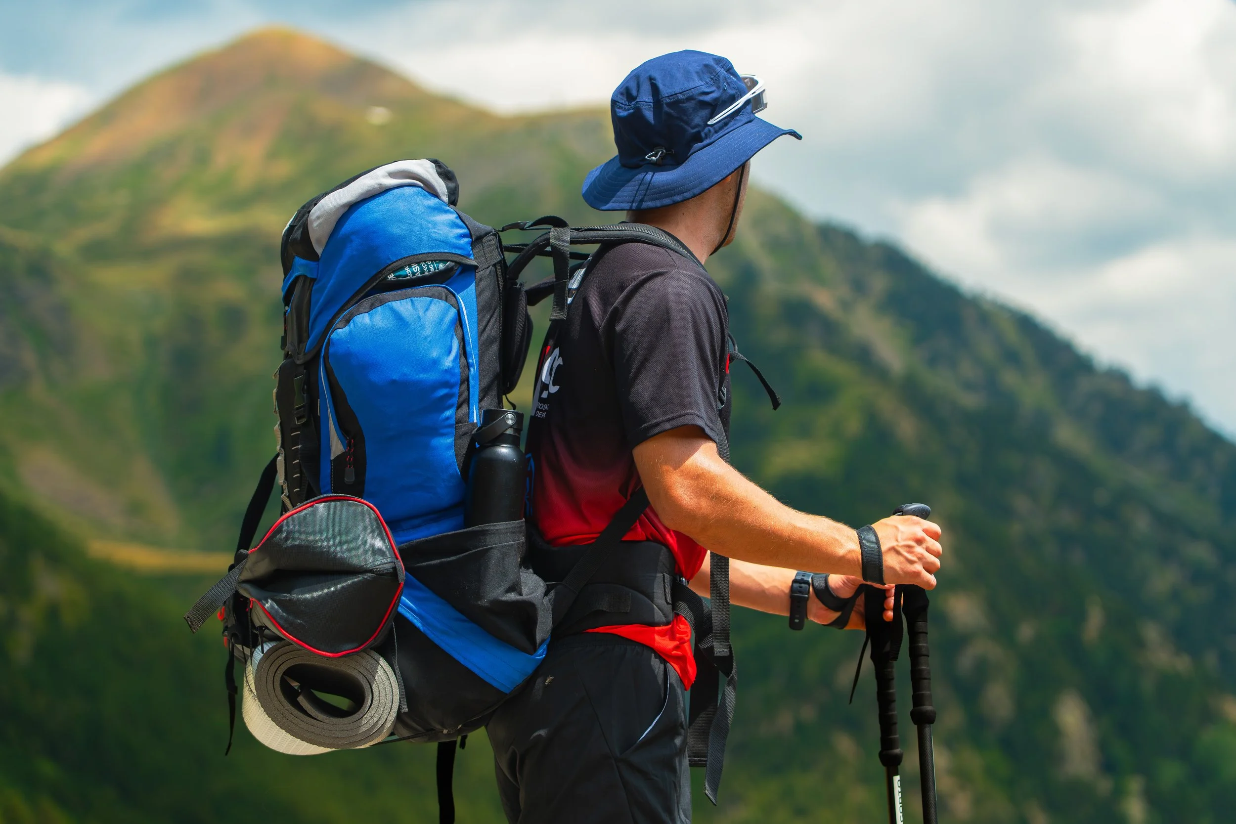 Persona de espaldas haciendo senderismo con mochila y sombrero en un paisaje montañoso.