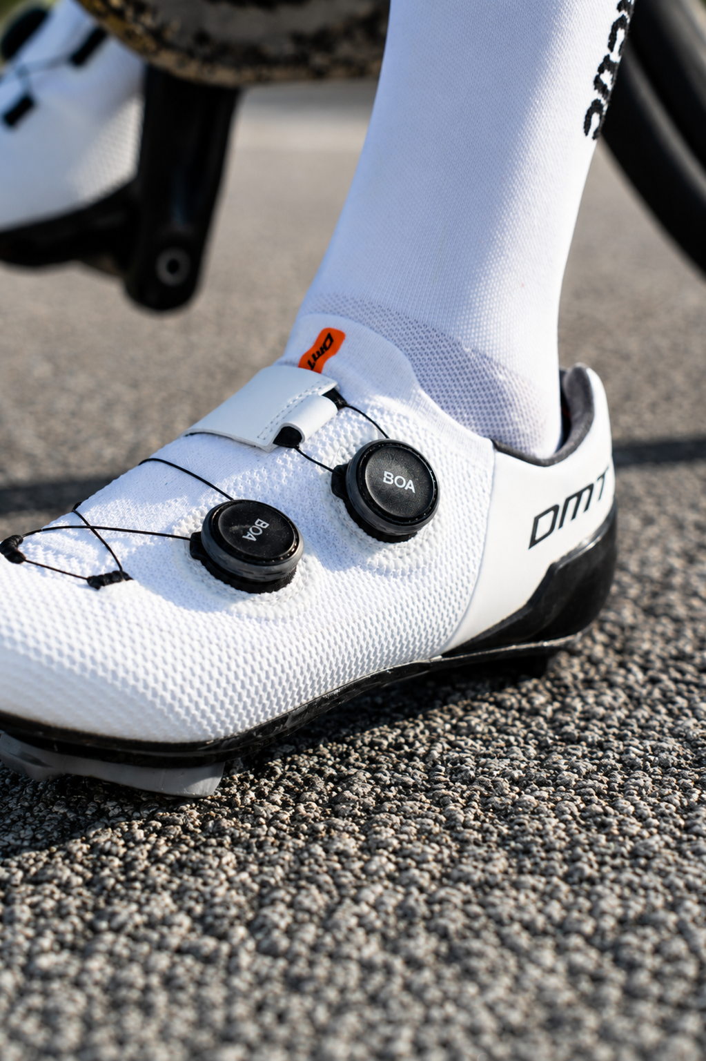 Zapatos de ciclismo blancos con calzado Shimano y accesorios BOA en una bicicleta, sobre un pavimento de carbono.