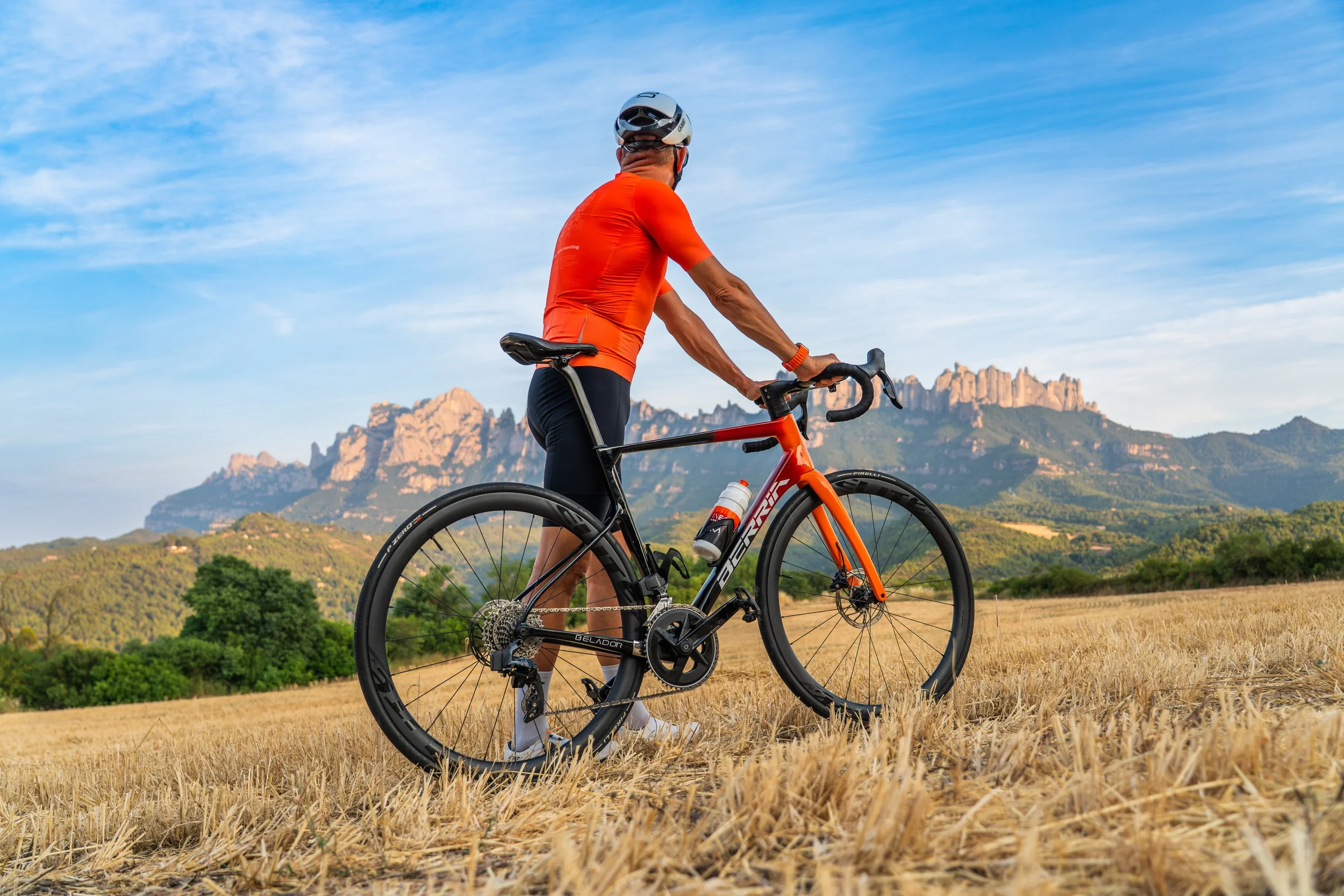 Persona con camiseta naranja y casco sobre bicicleta en campo con montañas al fondo.