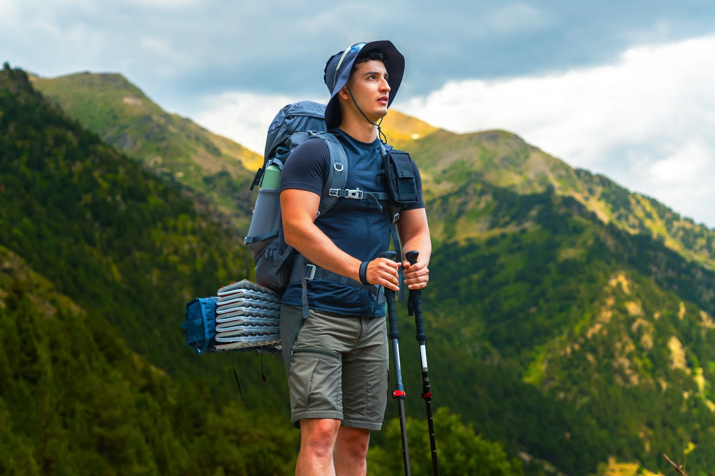 Joven hombre haciendo senderismo en la montaña, usando una gorra de ala ancha, una camiseta azul, pantalones cortos, con una mochila y bastones de senderismo, rodeado de un paisaje de montañas verdes y cielo nublado.