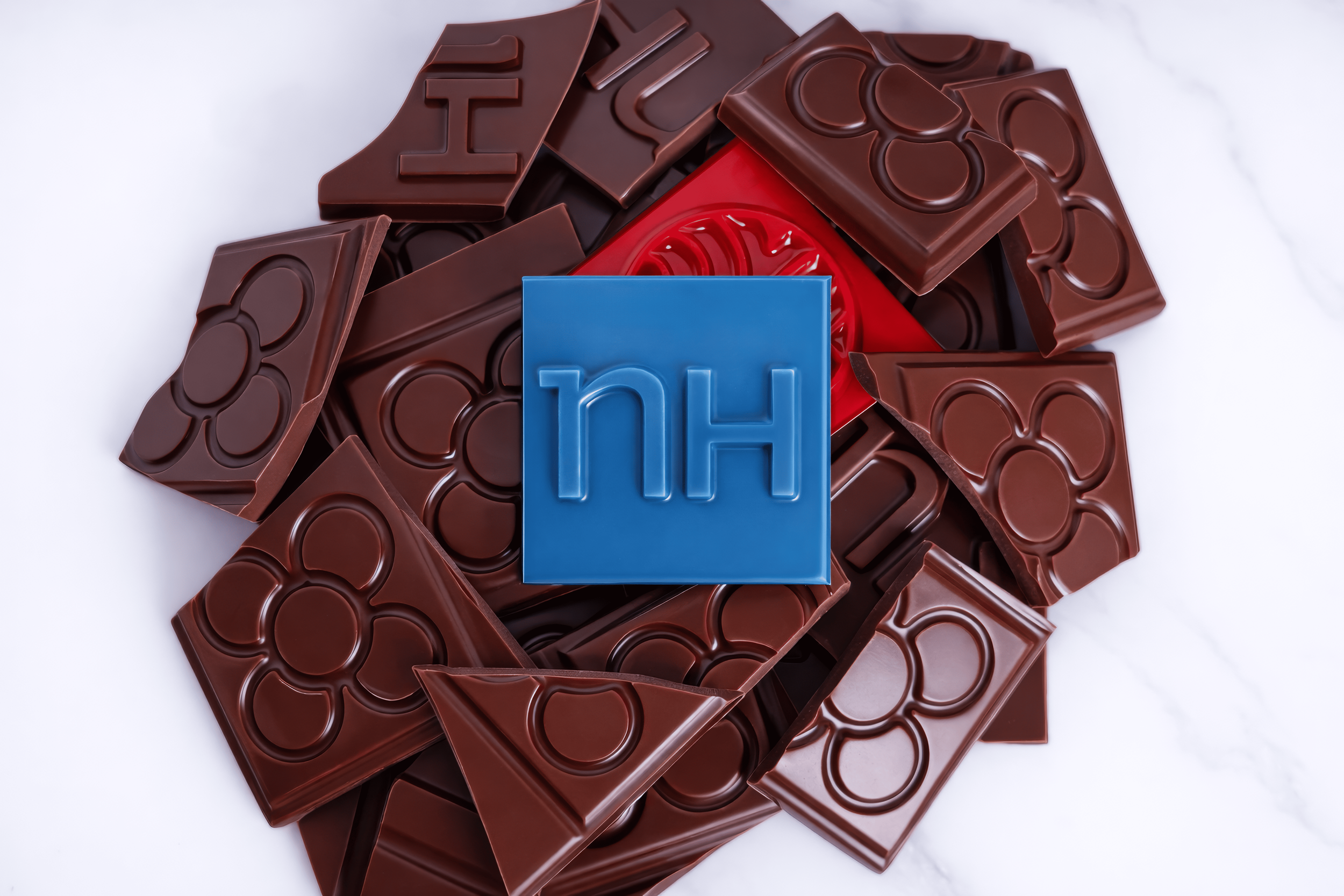 Varias piezas de chocolate con forma de flores, con una ficha azul en el centro que tiene el logo de 'nh'
