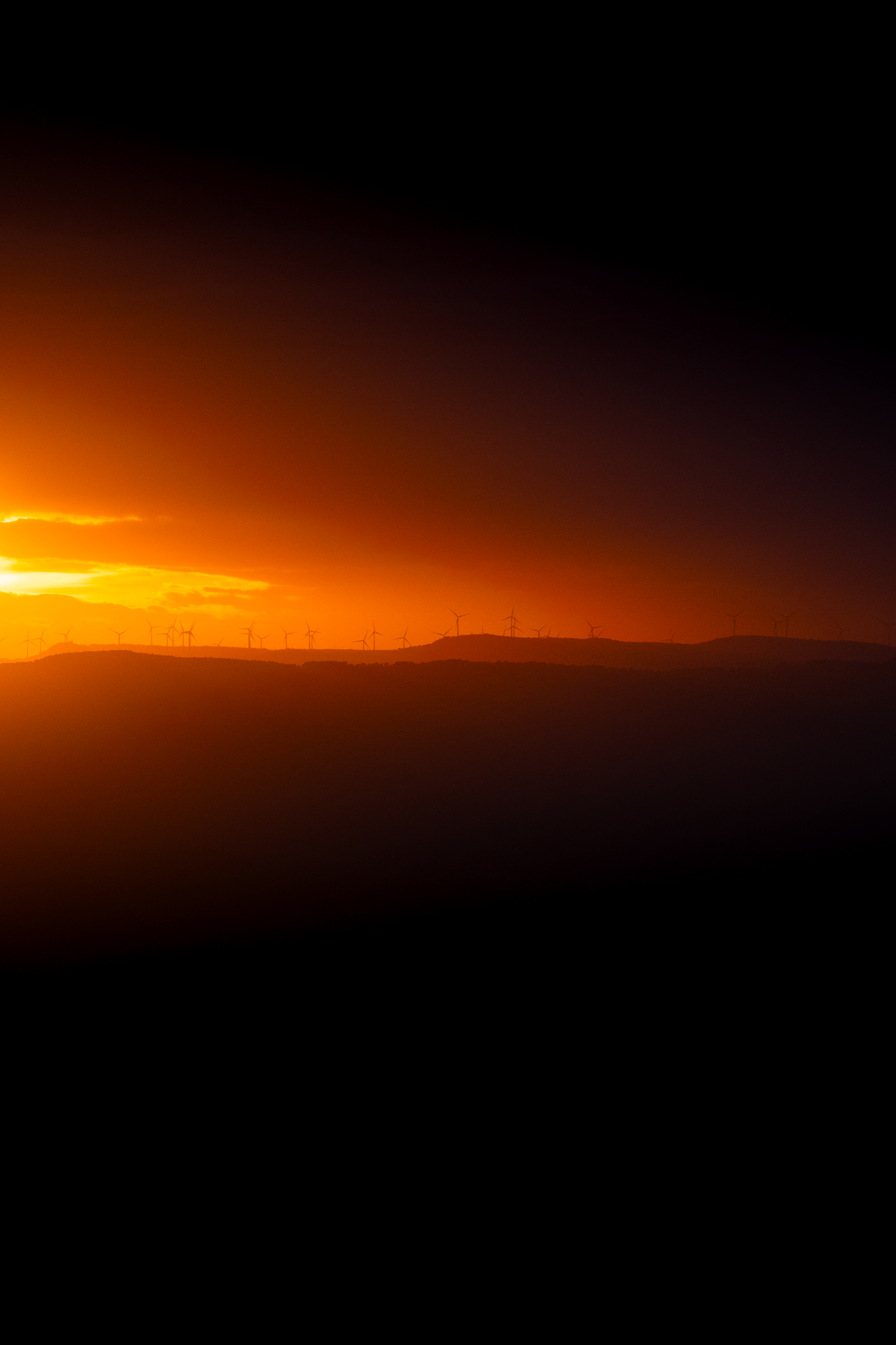 Atardecer con siluetas de molinos eólicos en un paisaje montañoso, cielo de tonos anaranjados y oscuros.