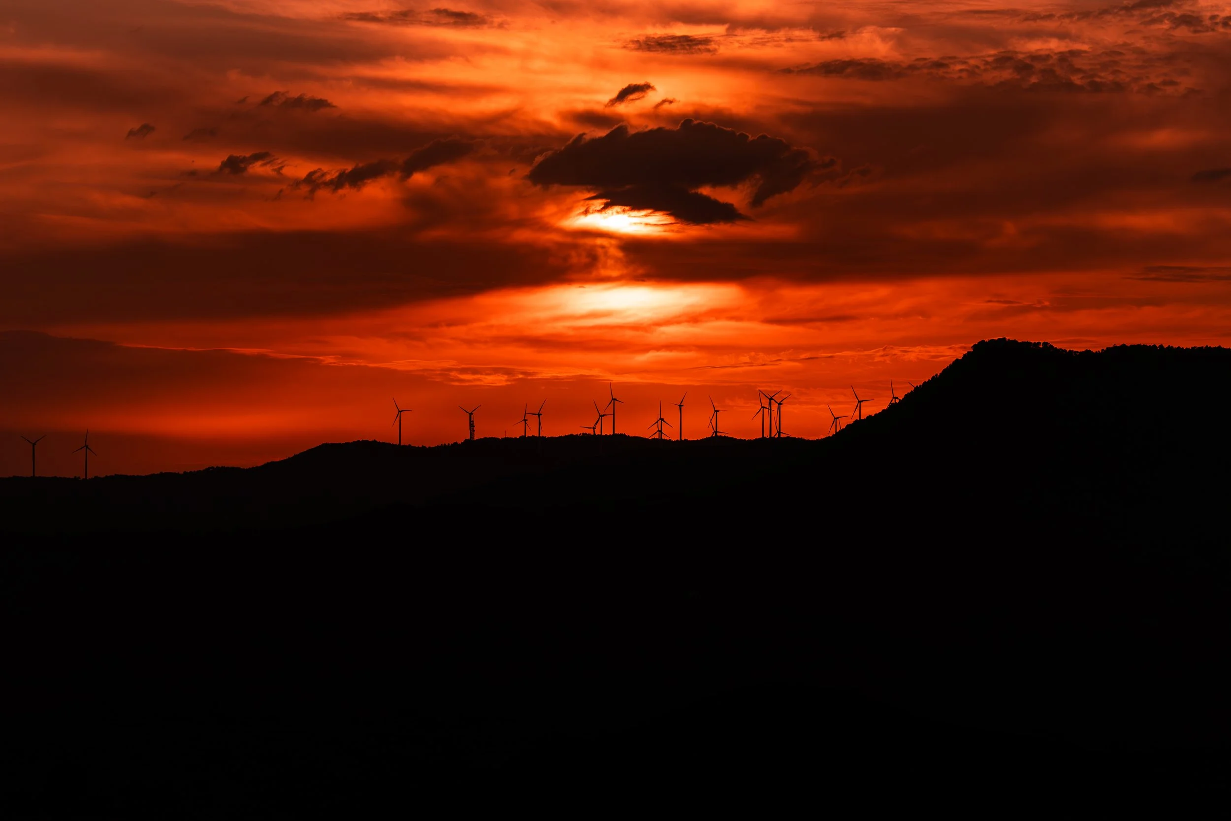 Atardecer con nubes oscuras y un cielo anaranjado, siluetas de aerogeneradores en un paisaje montañoso.