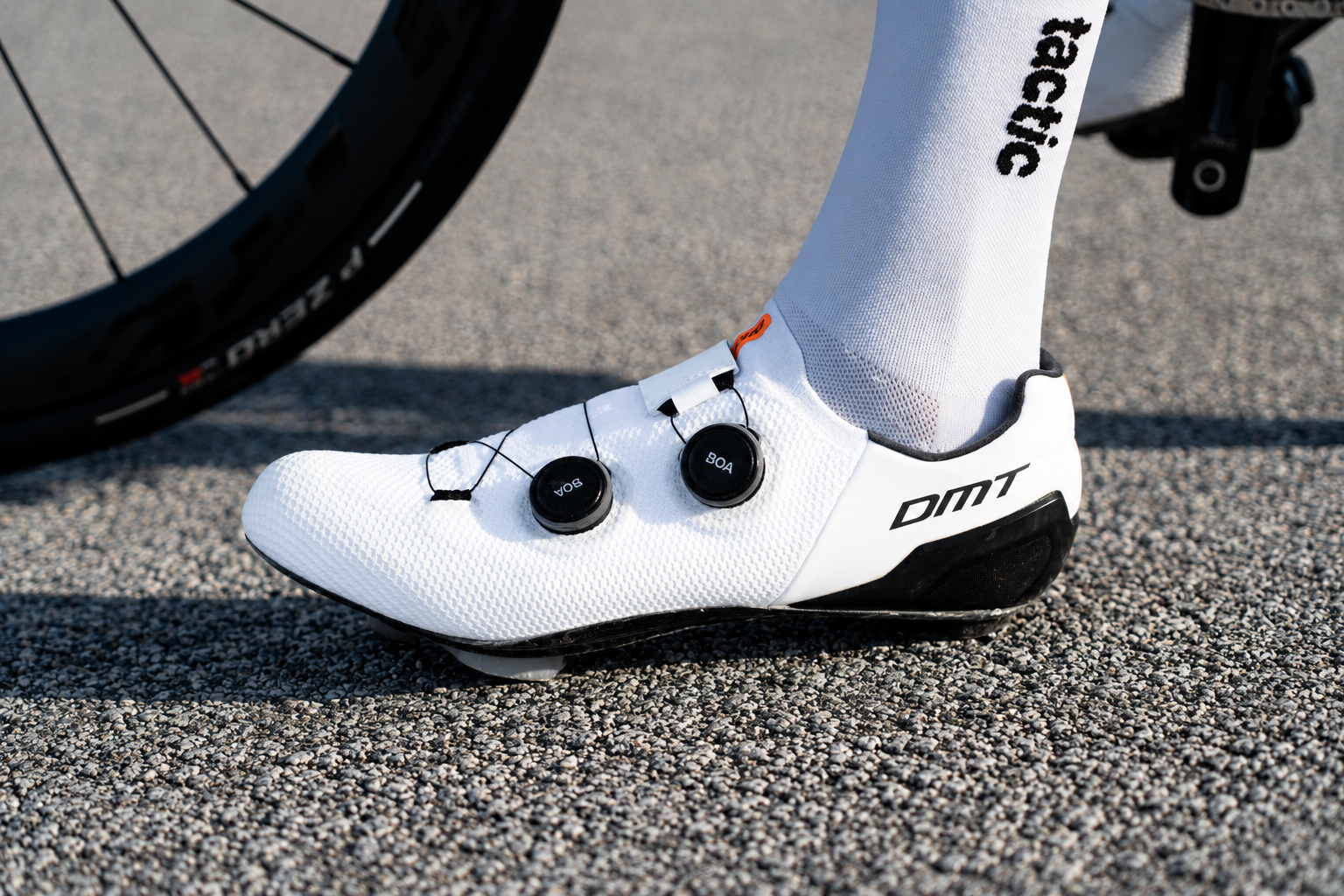 Zapatos de ciclismo blancos con calcomanías negras y un sistema de cierre con tuercas negras, usados por una persona en un camino de asfalto.