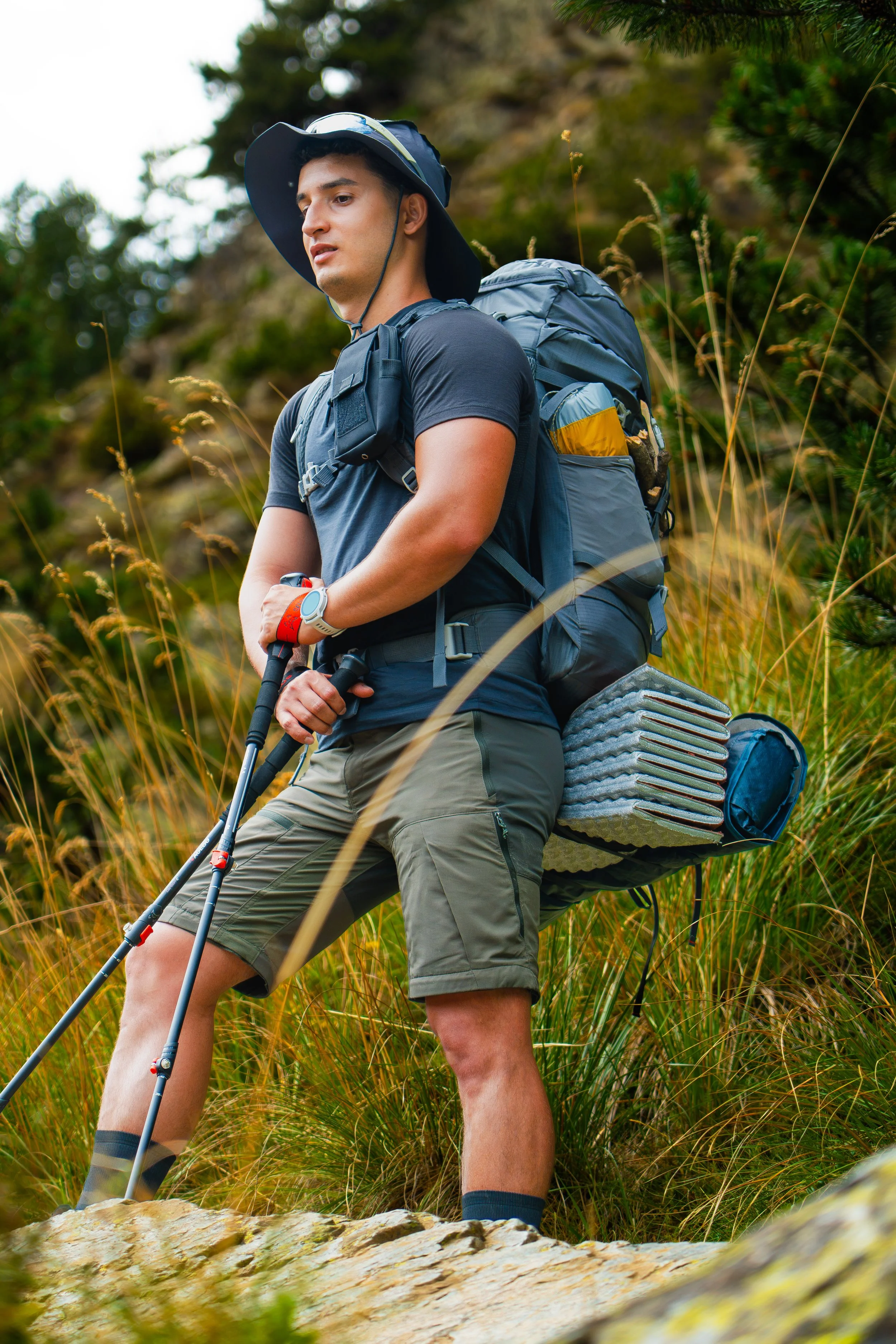 Joven hombre haciendo senderismo en un área natural, con mochila y bastones, en un entorno de vegetación y rocas.