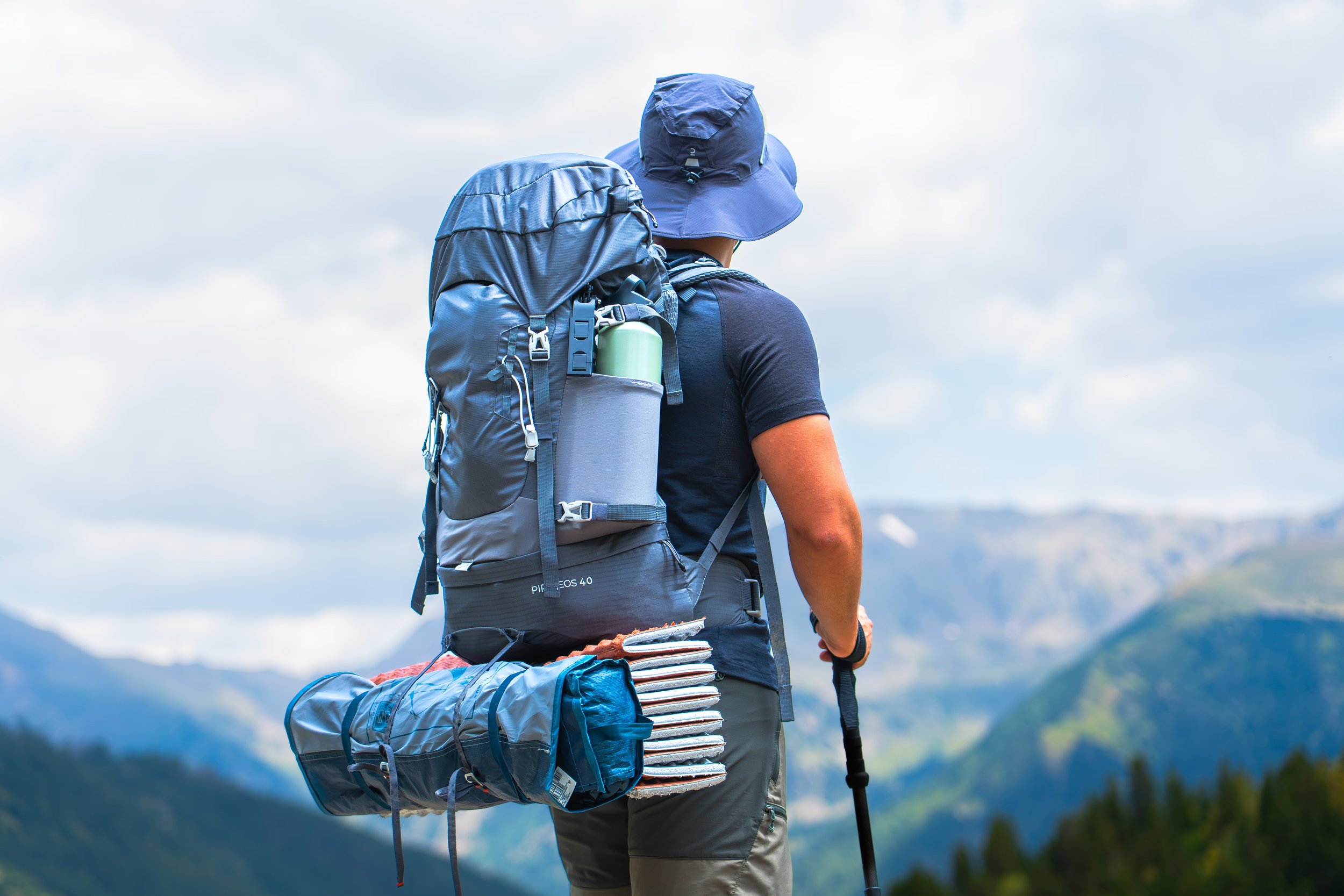 Persona con sombrero azul, mochila en la espalda, y caminando en un paisaje montañoso con cielo nublado.