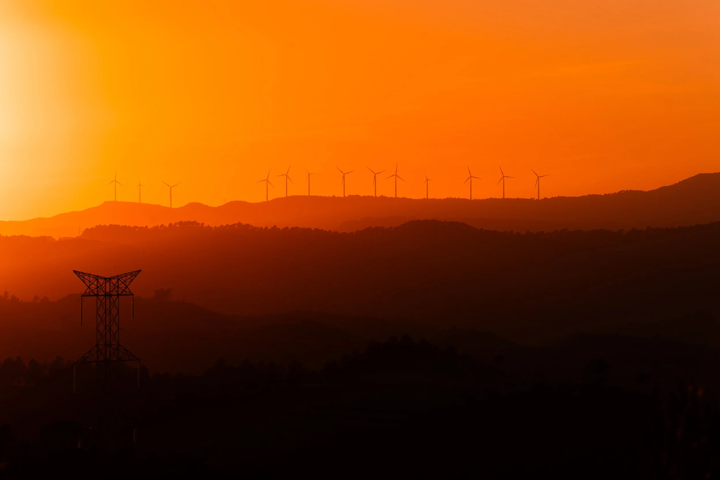 Paisaje con colinas y molinos de viento durante un atardecer con cielo naranja.